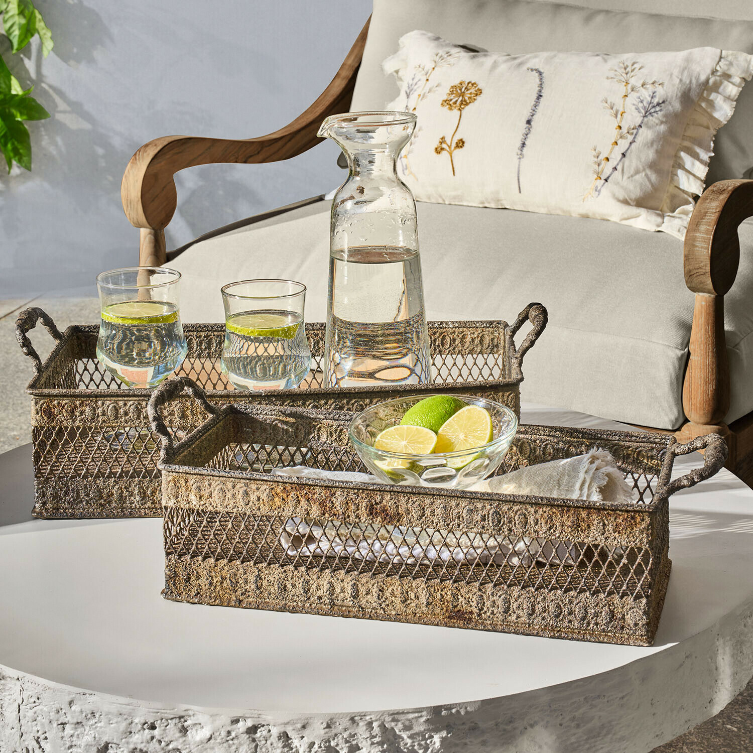 Dos bandejas metálicas rústicas con asas sobre mesa blanca con jarra y vasos de agua con lima