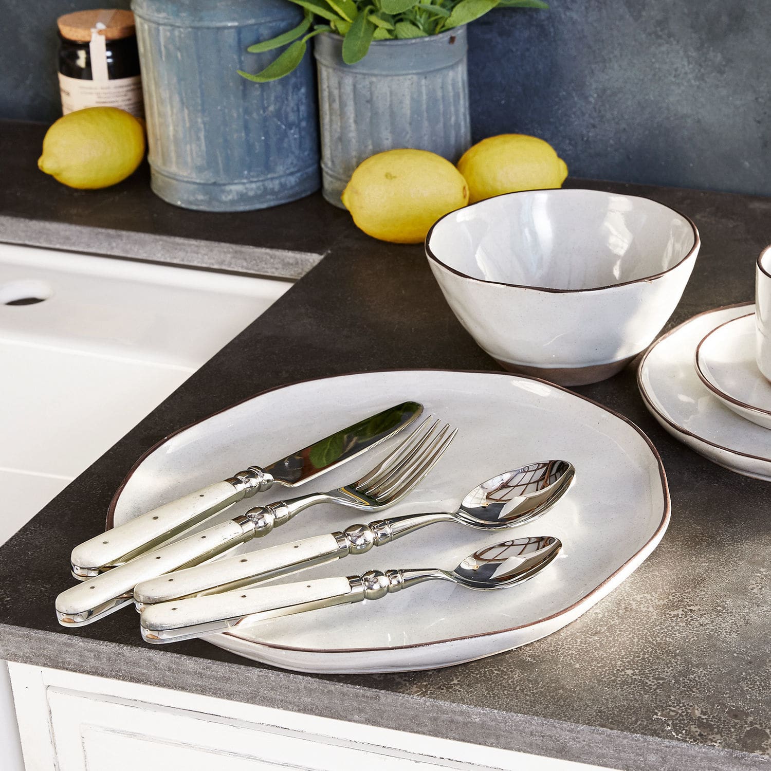 Set de 6 platos Biarré Juego de cubiertos con mango blanco sobre plato y cuenco de cerámica en encimera con limones