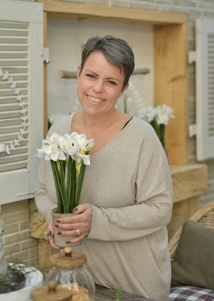 Mujer sonriente con suéter beige sosteniendo maceta con flores blancas en ambiente natural de Pascua