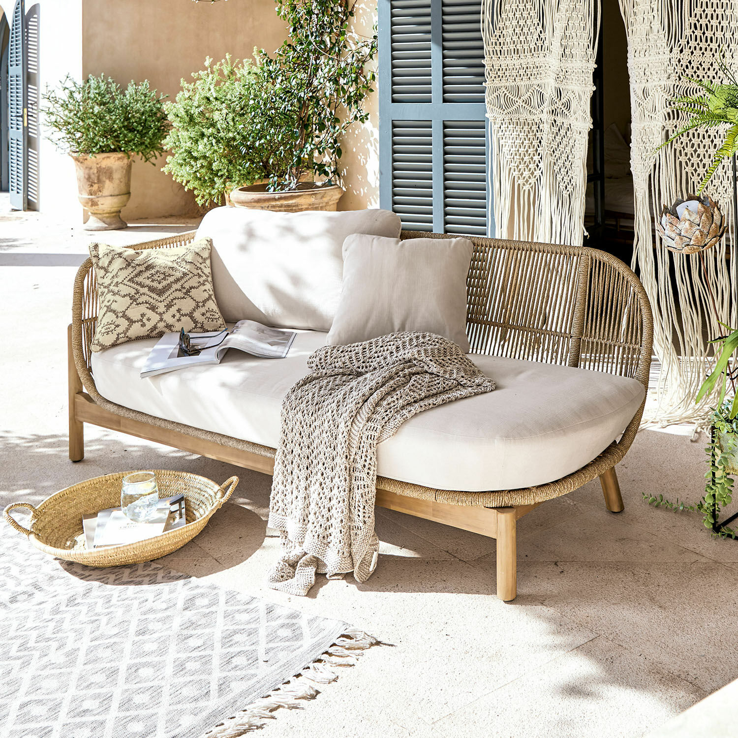 Sofá de ratán y madera con cojines crema y manta en terraza luminosa con plantas