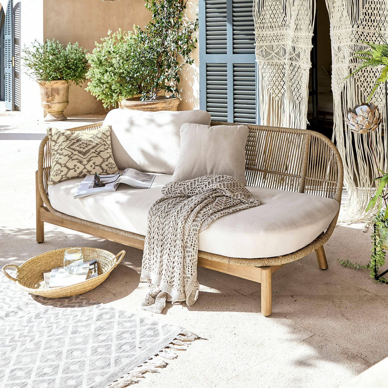 Chaise longue FSC®-100% Belvidere Sofá de ratán y madera con cojines crema y manta en terraza luminosa con plantas