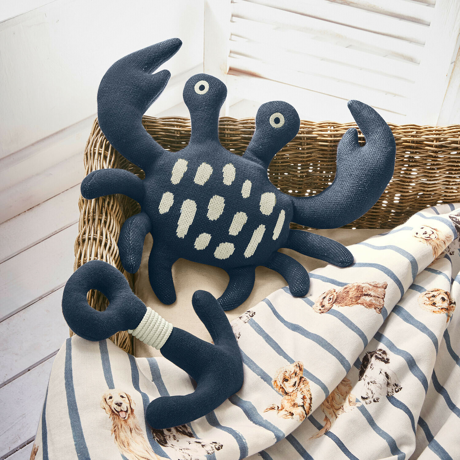 Juguete de peluche azul marino en forma de cangrejo y ancla sobre manta a rayas con diseño de perros