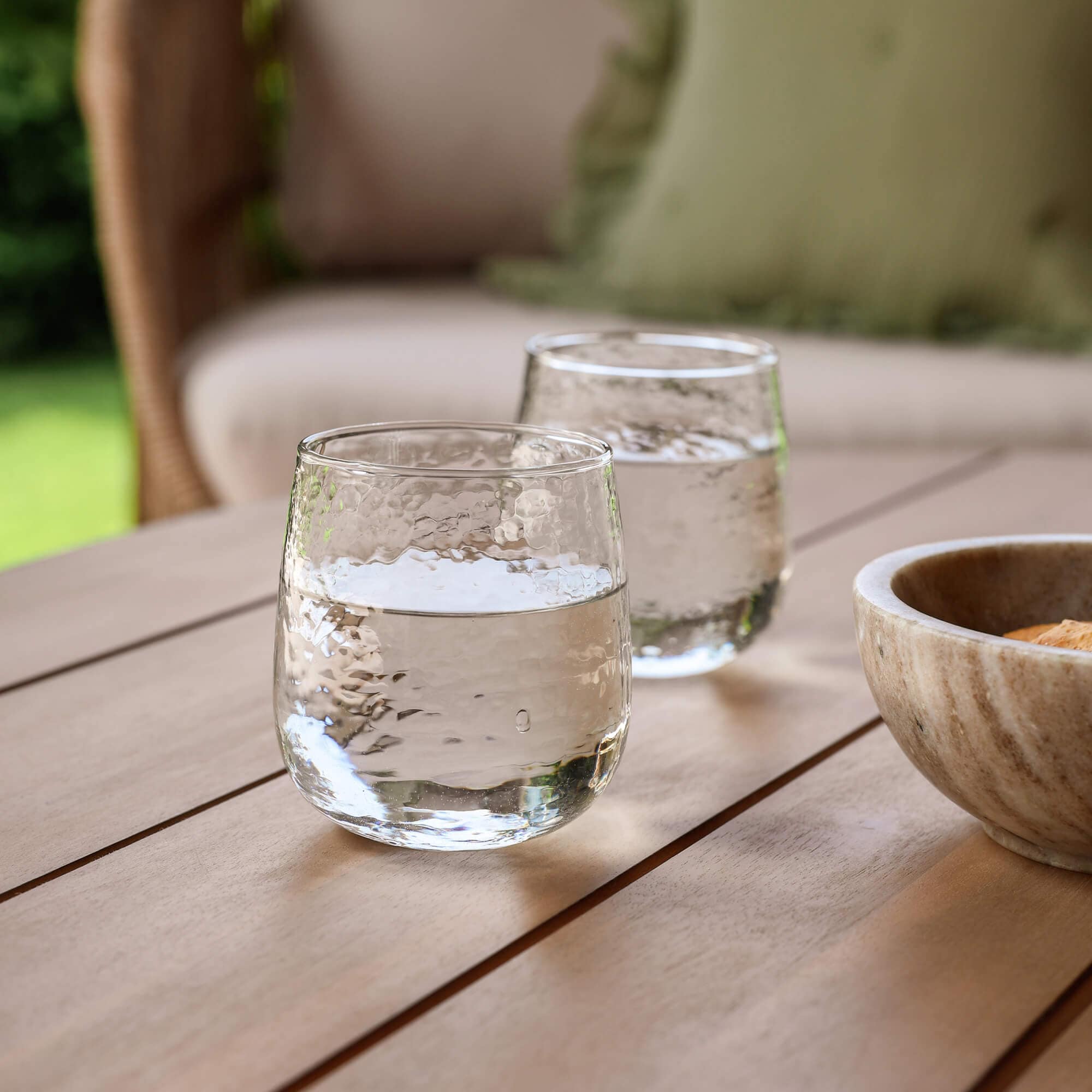 Dos vasos transparentes con agua sobre una mesa de madera en un jardín junto a un cuenco de piedra