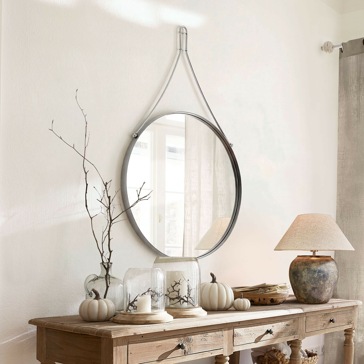 Espejo Célivaux Espejo redondo colgado con correa metálica plateada sobre consola de madera con decoración otoñal
