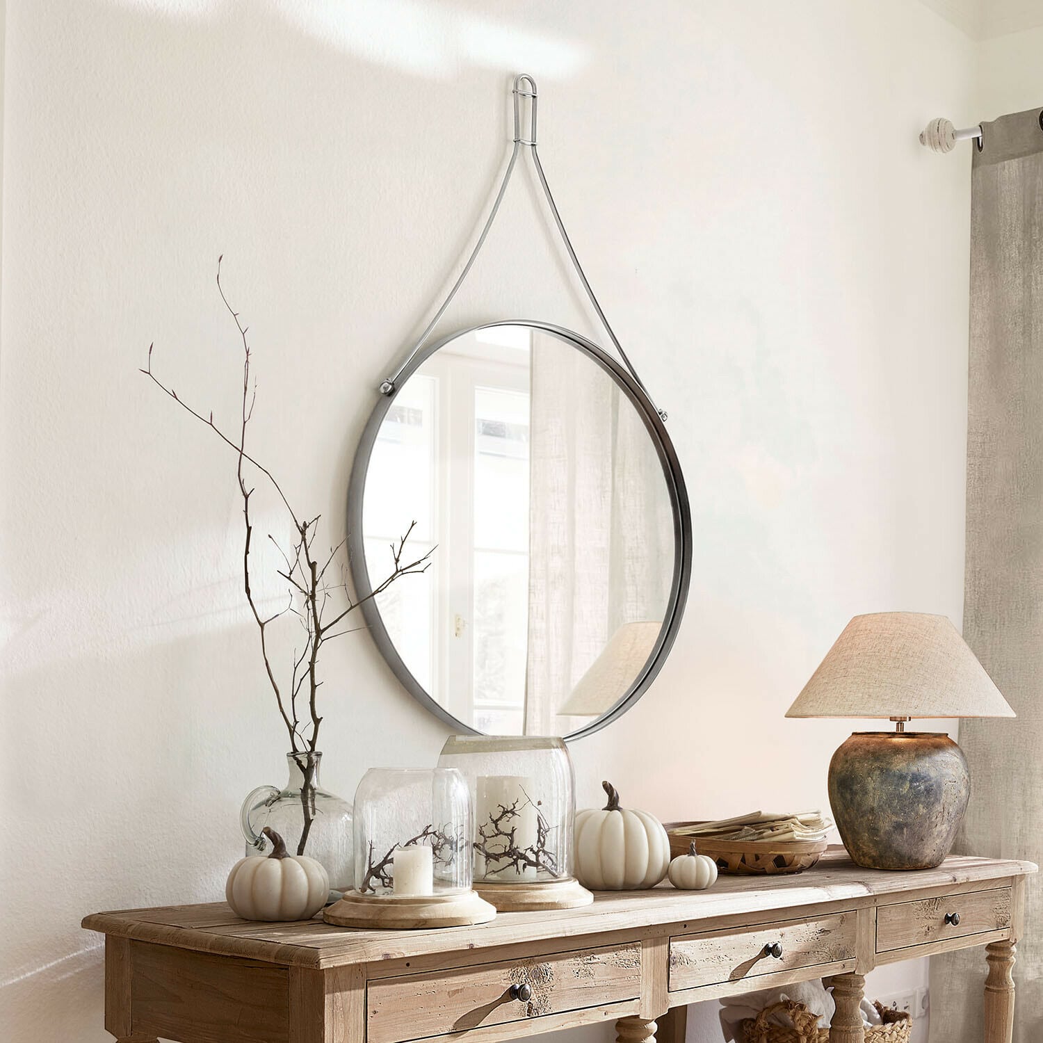 Espejo Célivaux Espejo redondo colgado con correa metálica plateada sobre consola de madera con decoración otoñal