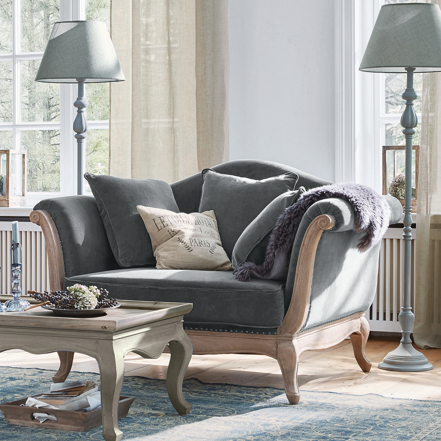 Sillón Sumerset Sofá gris de terciopelo con marco de madera y cojines en sala luminosa con lámparas de pie