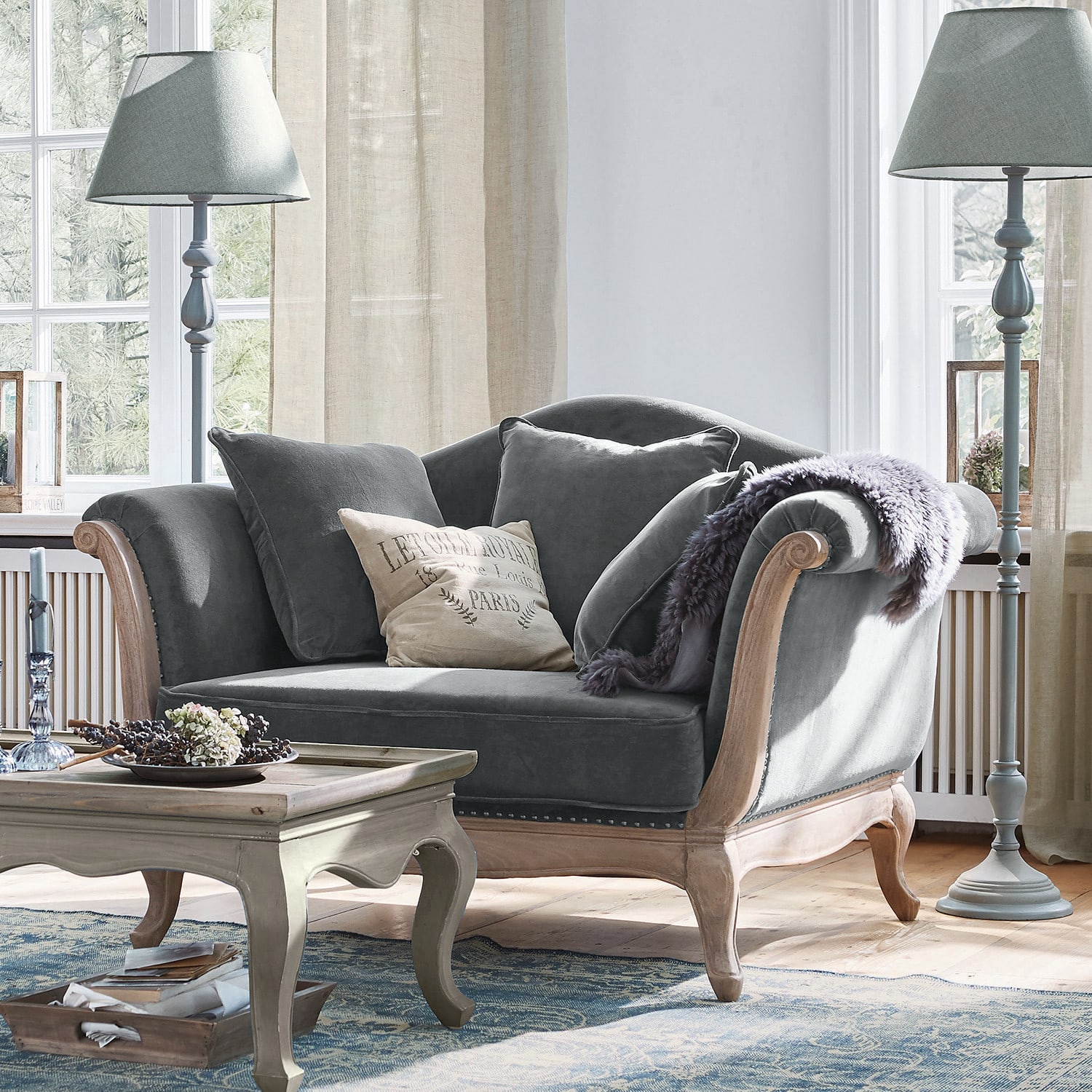 Sillón Sumerset Sofá gris de terciopelo con marco de madera y cojines en sala luminosa con lámparas de pie