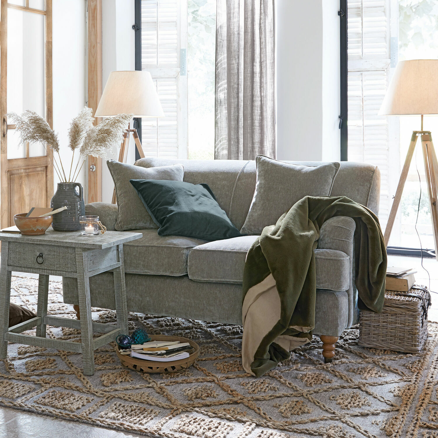 Sofá gris con cojines suaves y manta verde en sala luminosa con lámparas de madera y alfombra