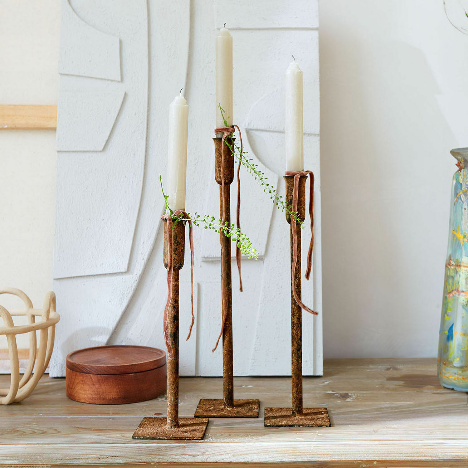Set de 3 candeleros Viverto Tres candelabros de metal oxidado con velas blancas y ramas verdes decorativas sobre mesa de madera