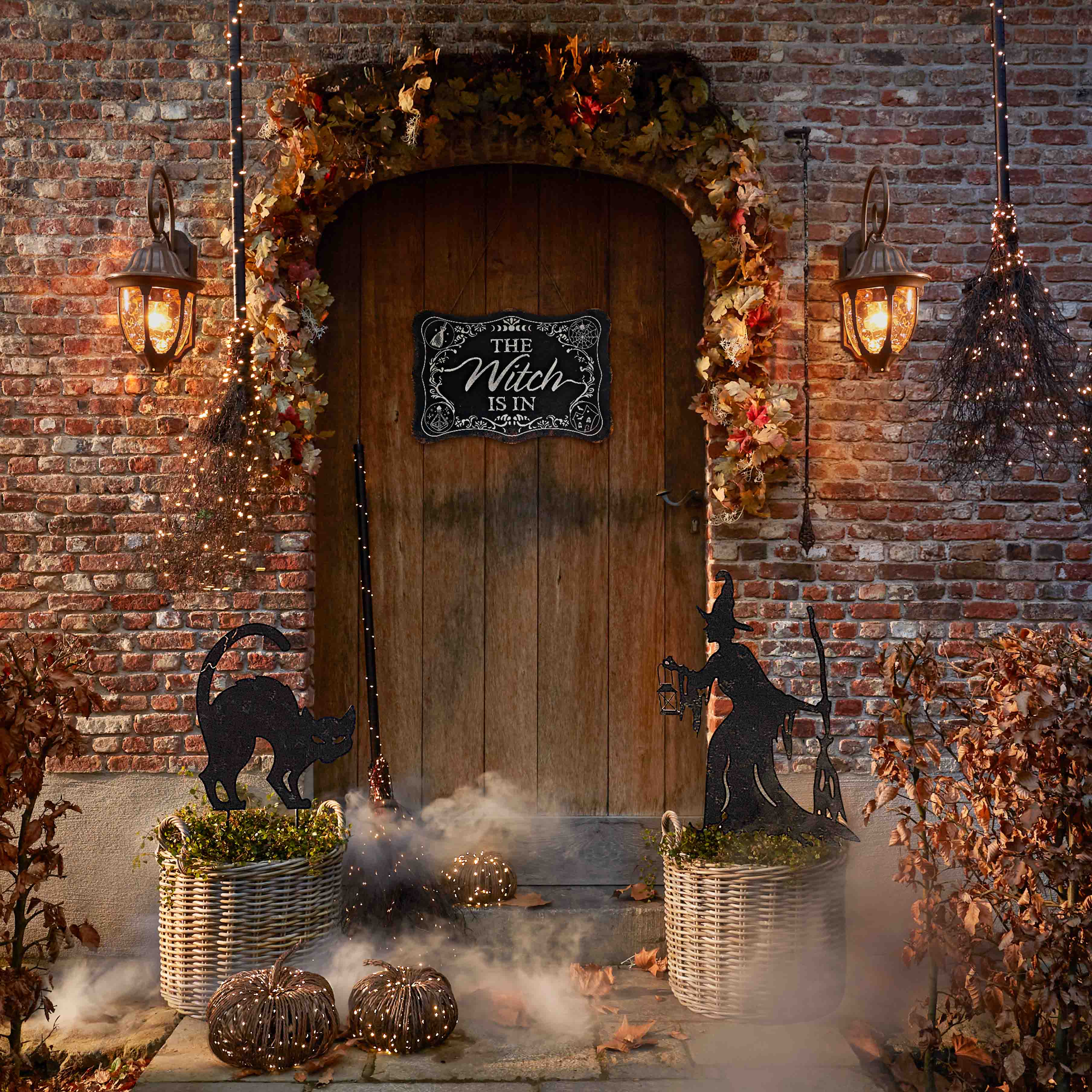 Decoración de Halloween con gato negro, bruja y faroles frente a puerta de madera y pared de ladrillo