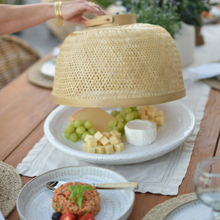 Campana de bambú natural cubre queso y uvas verdes en plato blanco sobre mesa de madera