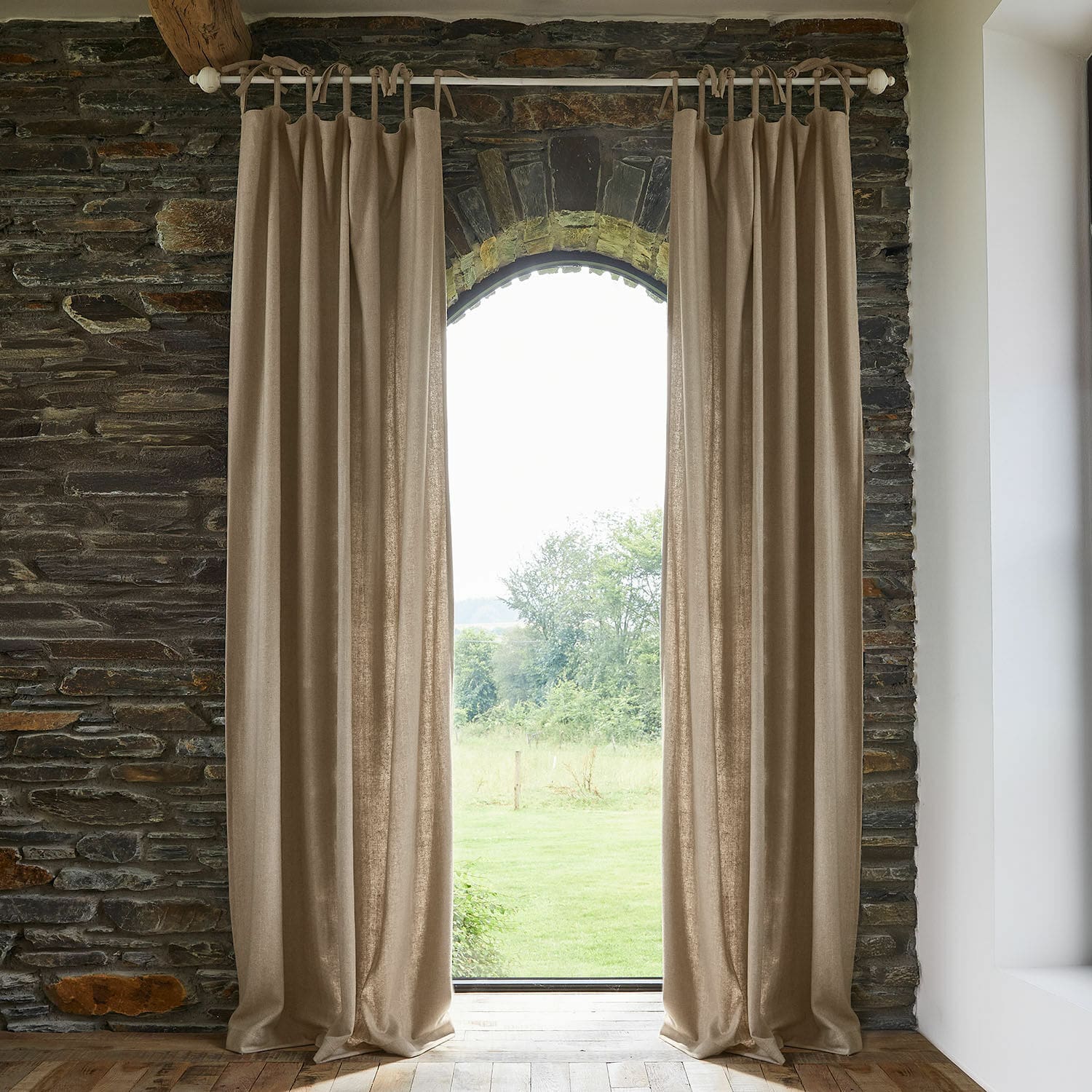 Cortina Seleneia Cortinas beige de tela natural colgadas en barra en interior de piedra con vista al jardín