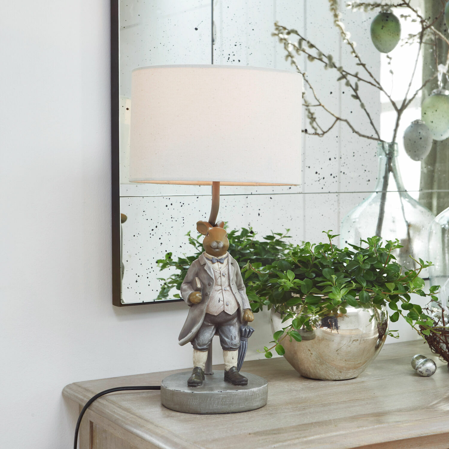 Lámpara de mesa Cerni Lámpara de mesa con figura de conejo vestido de gris y pantalla blanca sobre mueble de madera