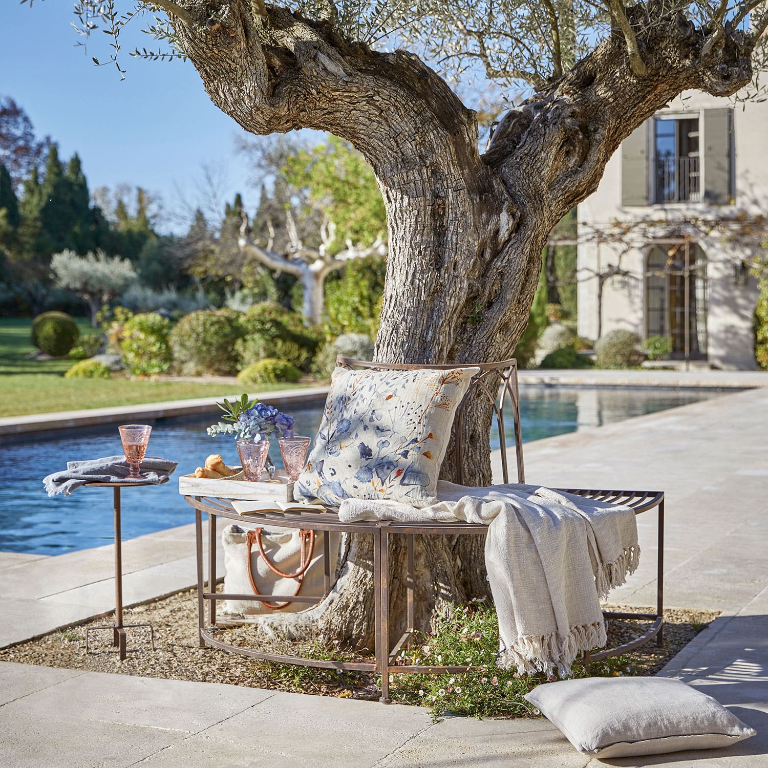 Banco de metal alrededor de un árbol con cojín y manta junto a una piscina en jardín