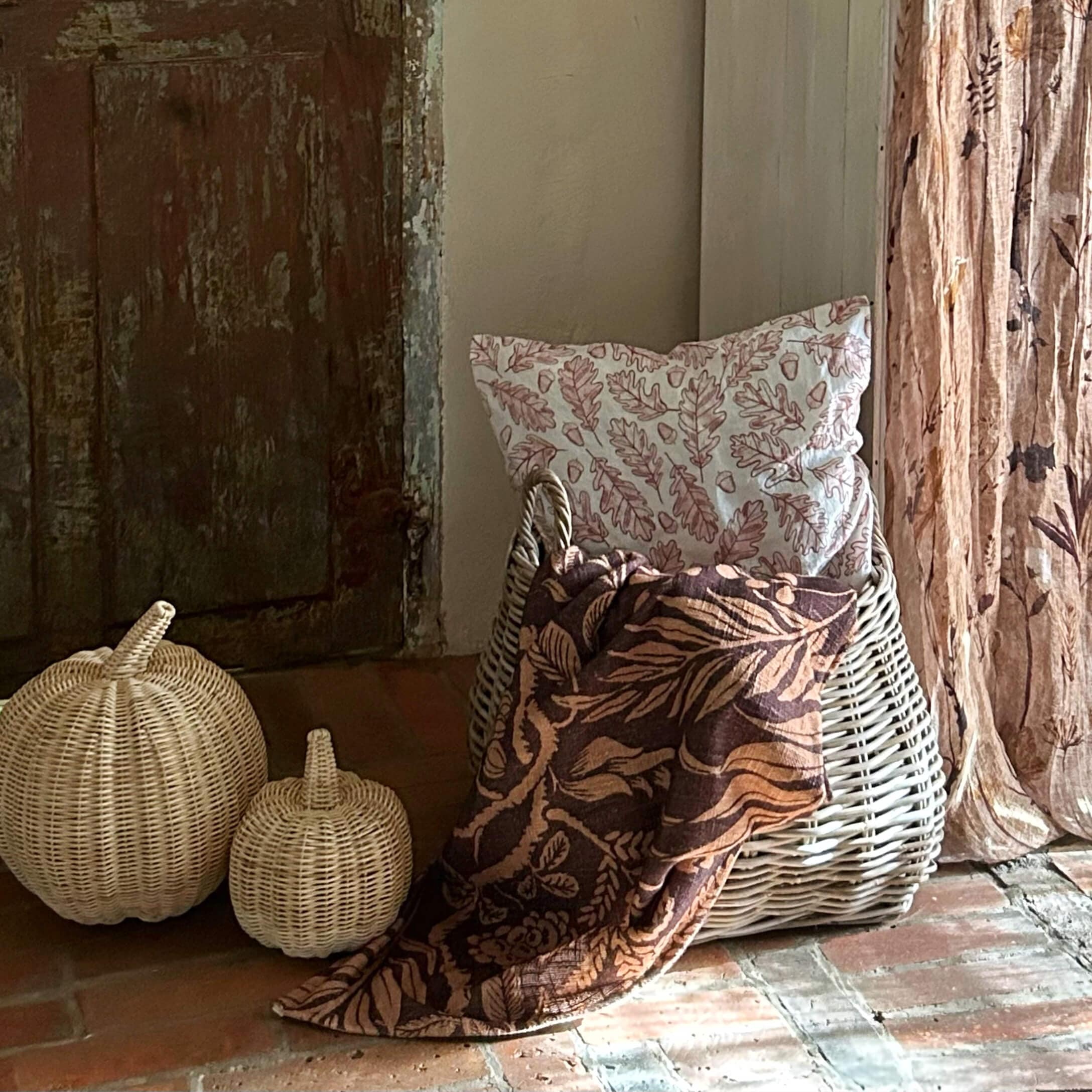 Cesta de mimbre blanca con cojín de hojas y manta marrón con motivos florales sobre suelo de ladrillo