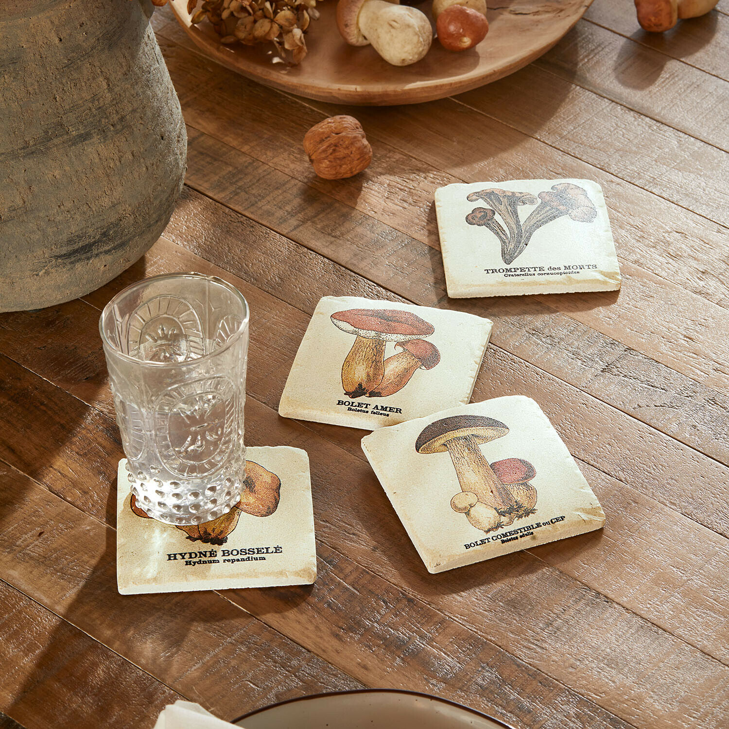 Set de 4 posavasos Jandoria Posavasos con ilustraciones de setas sobre mesa de madera con vaso transparente y nueces