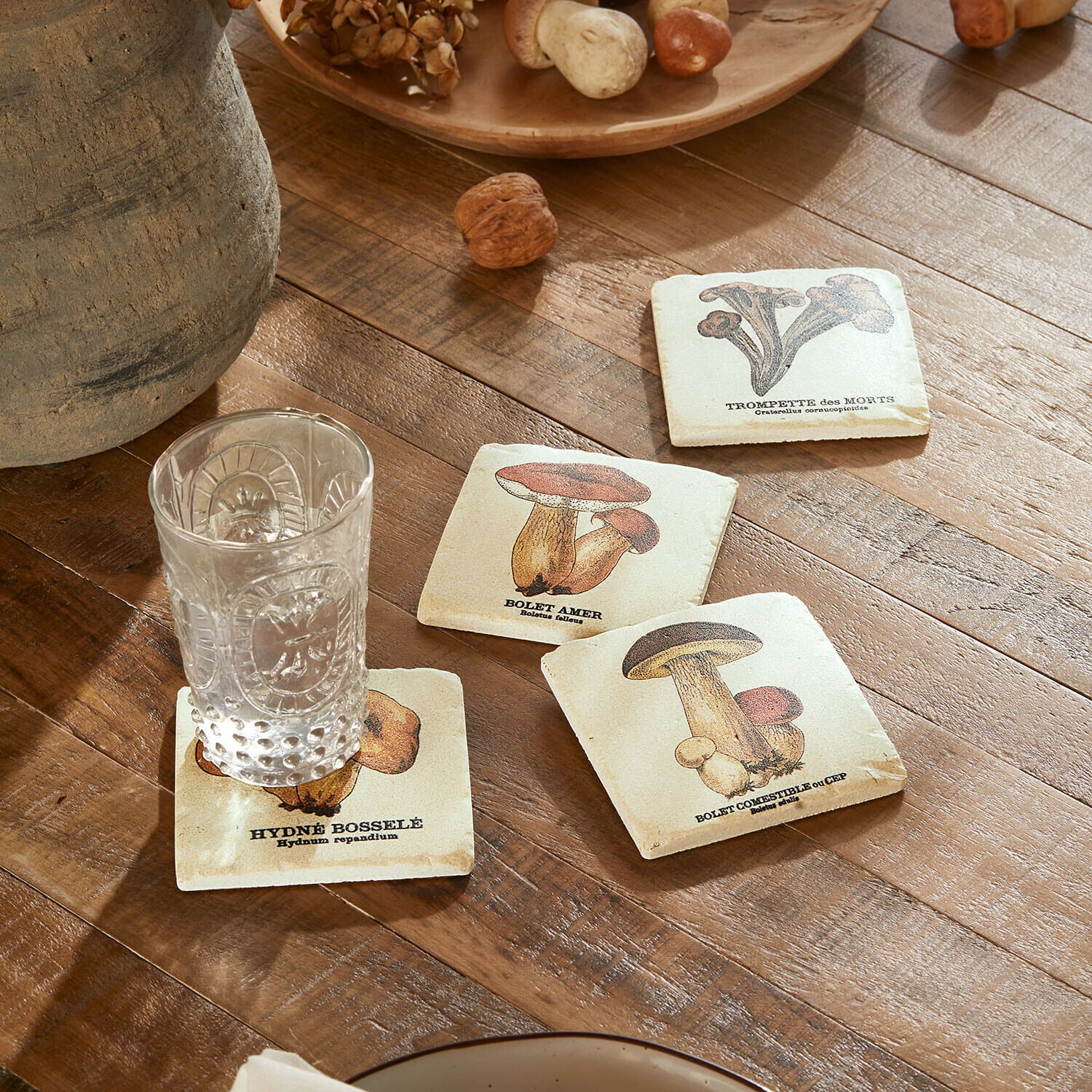 Set de 4 posavasos Jandoria Posavasos con ilustraciones de setas sobre mesa de madera con vaso transparente y nueces