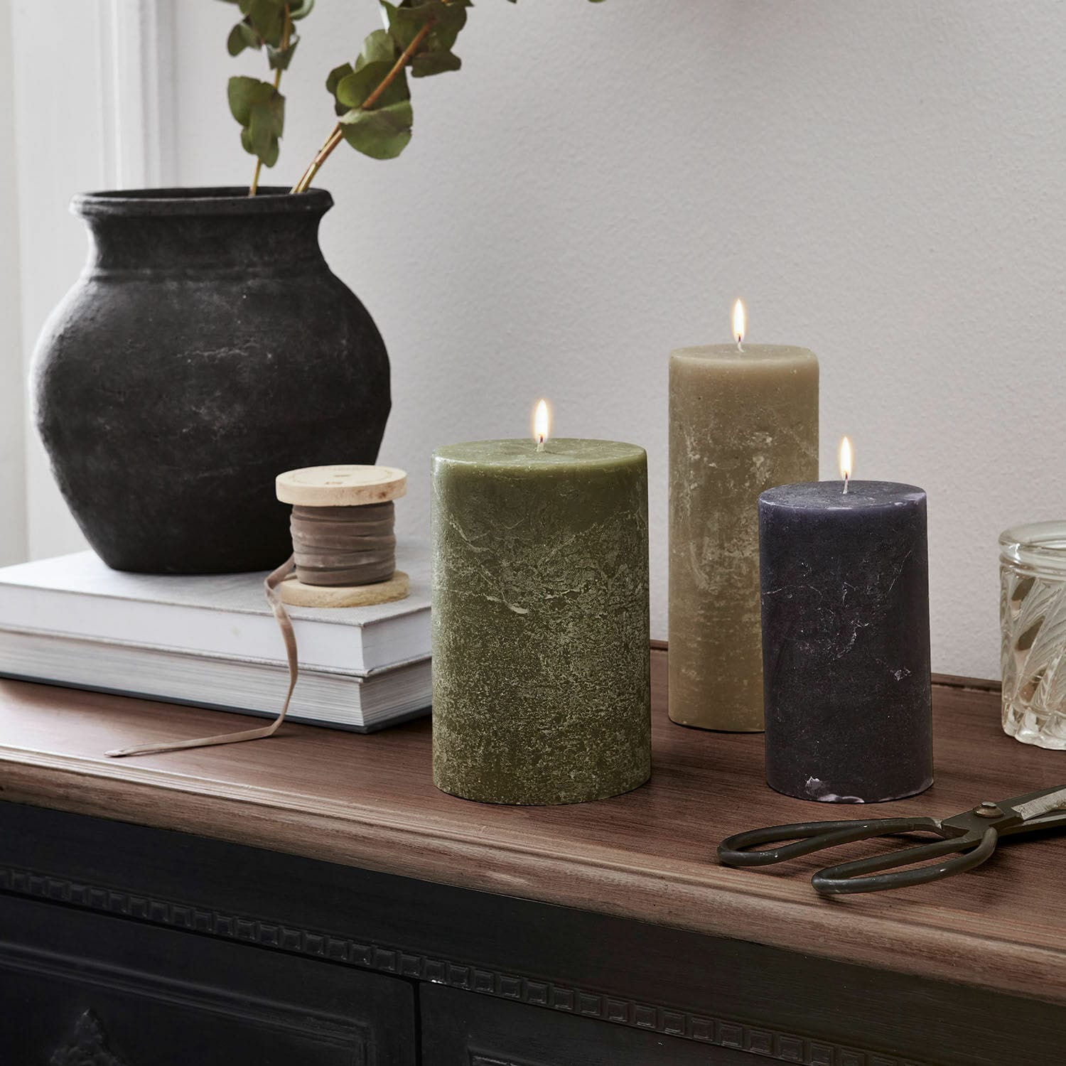 Set de 3 velas Pretin Tres velas encendidas en verde, beige y negro sobre superficie de madera con decoraciones