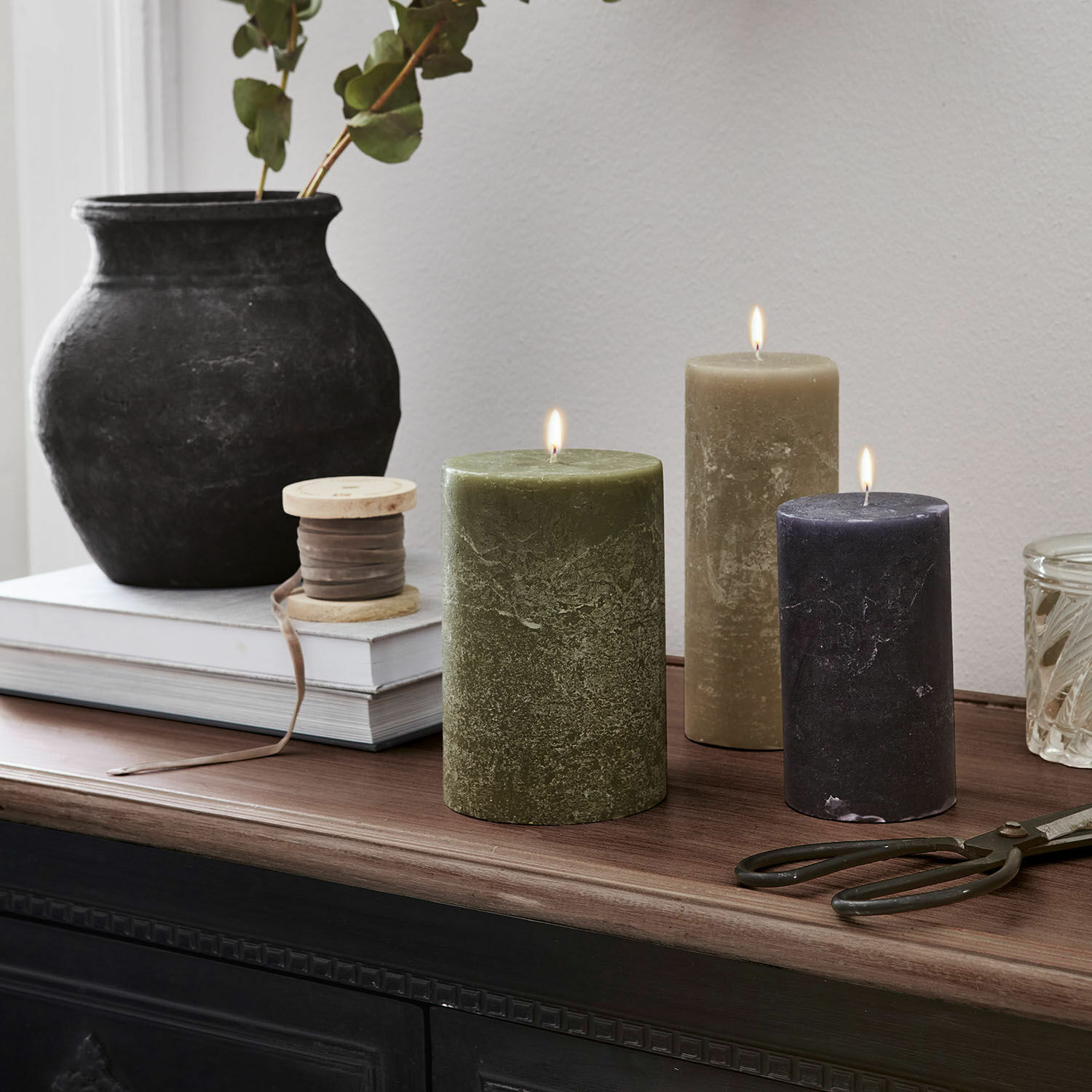Set de 3 velas Pretin Tres velas encendidas en verde, beige y negro sobre superficie de madera con decoraciones
