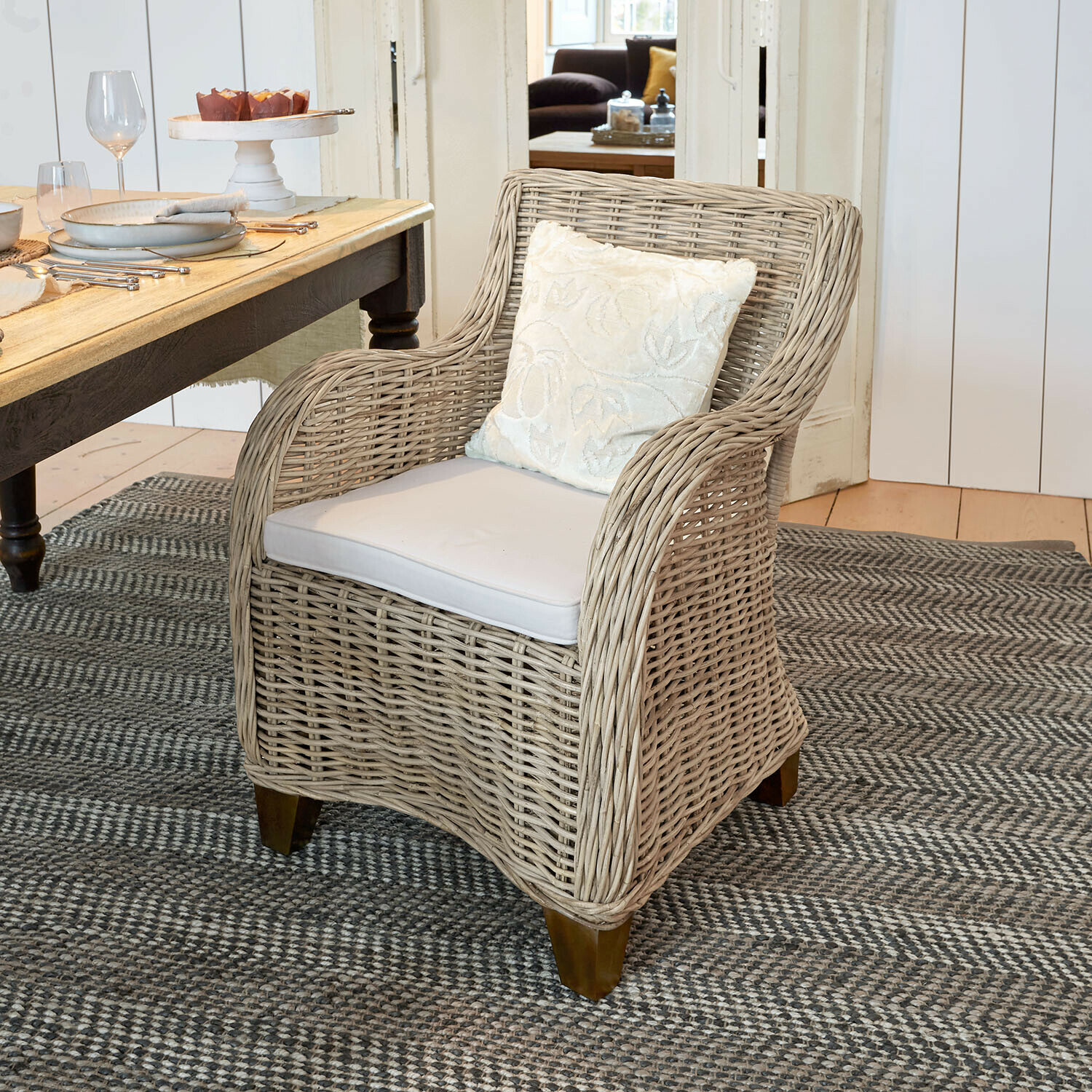 Silla Penrosse Sillón de mimbre beige con cojín blanco sobre alfombra gris en comedor