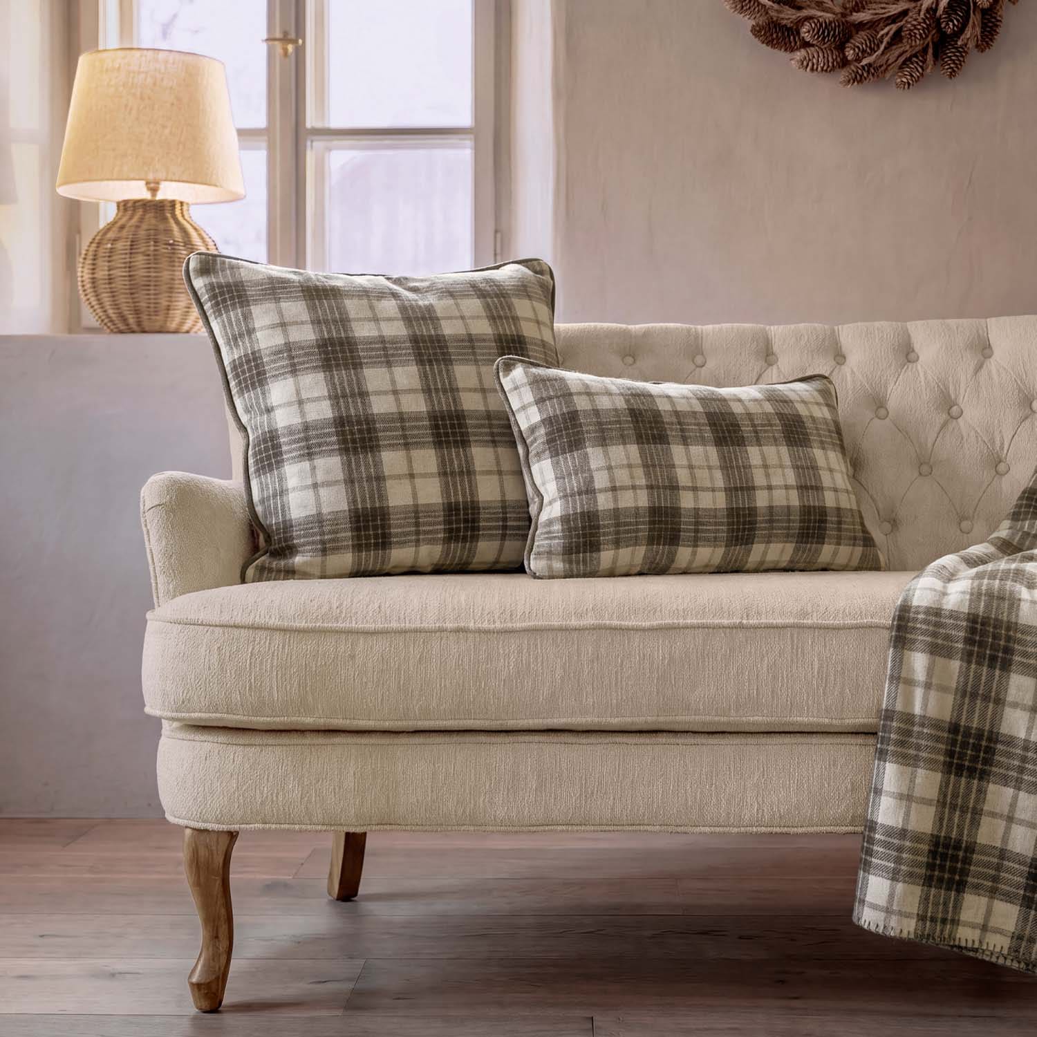 Sofá beige con dos cojines a cuadros en tonos grises y crema, lámpara de mimbre junto a ventana