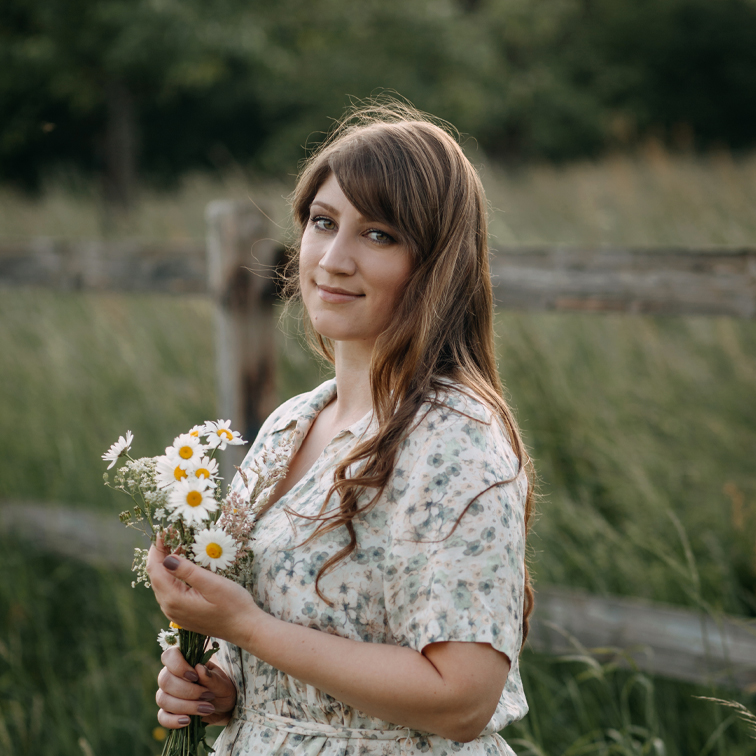 Mujer con vestido floral sosteniendo ramo de margaritas frente a campo verde y cerca de madera