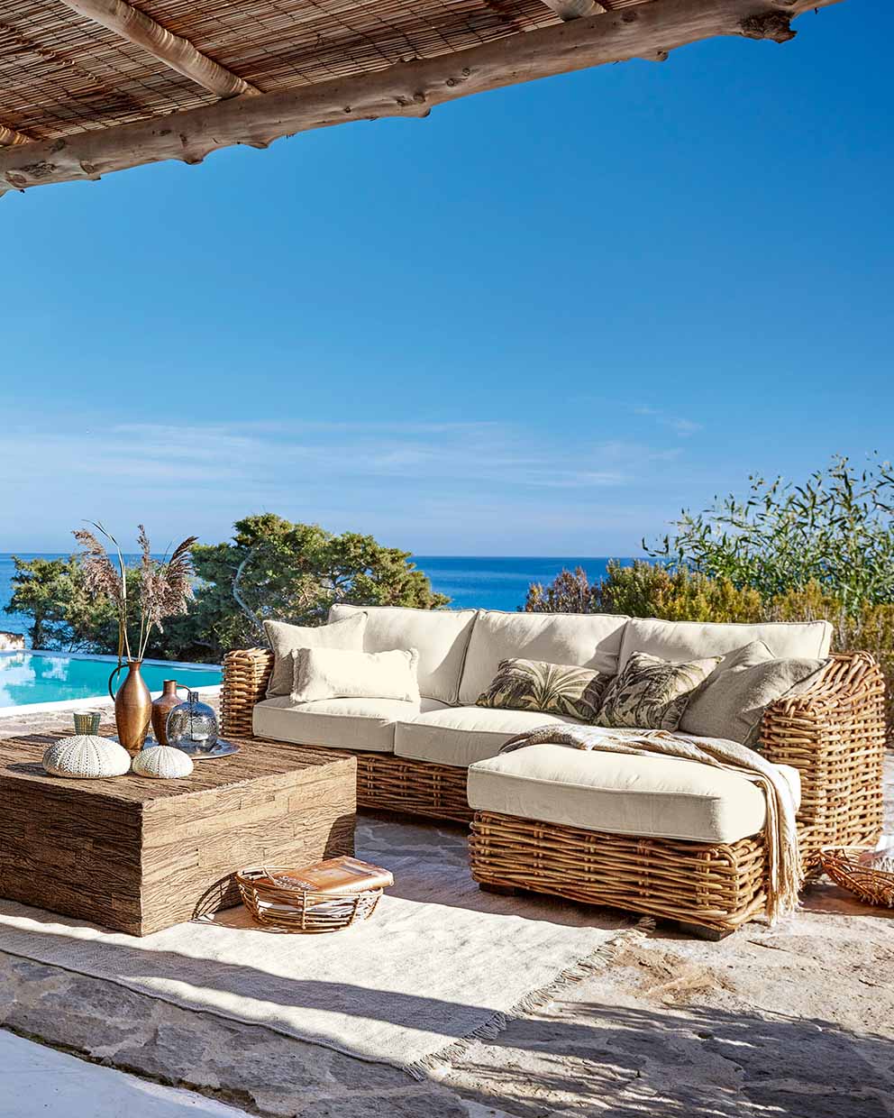 Conjunto de muebles de jardín de ratán con cojines crema y mesa de madera en terraza con vistas al mar