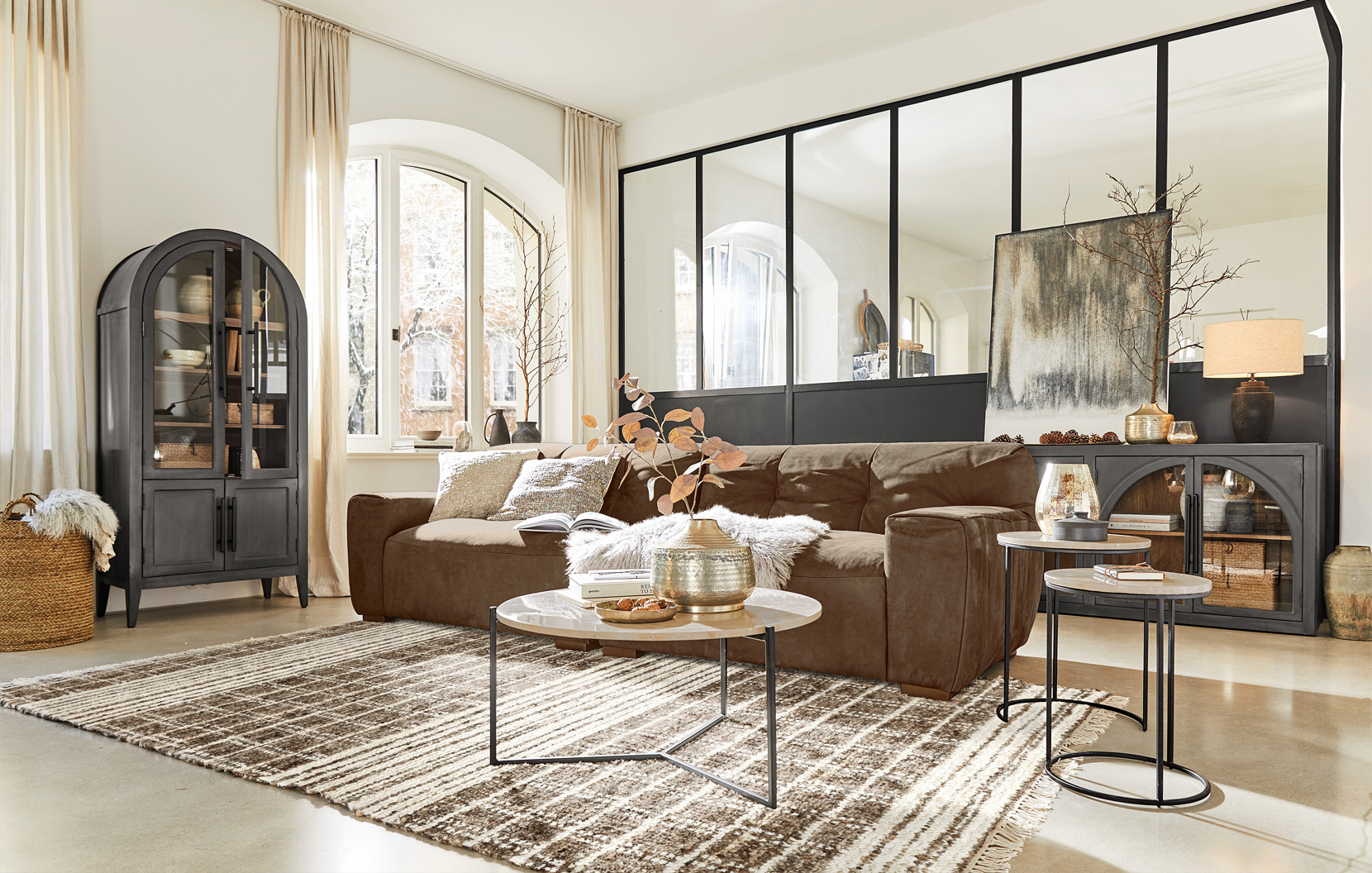 Salón acogedor con sofá marrón, mesas auxiliares metálicas y vitrina industrial negra con cristal