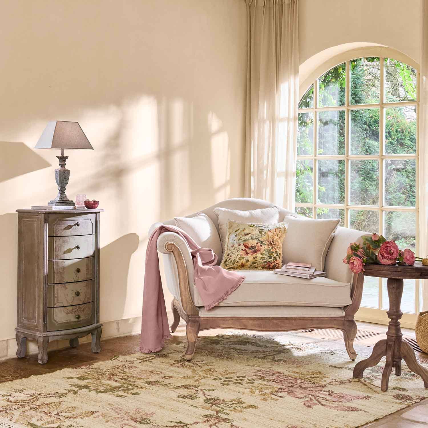 Sofá crema con marco de madera, cojín floral y manta rosa en sala luminosa con ventana grande