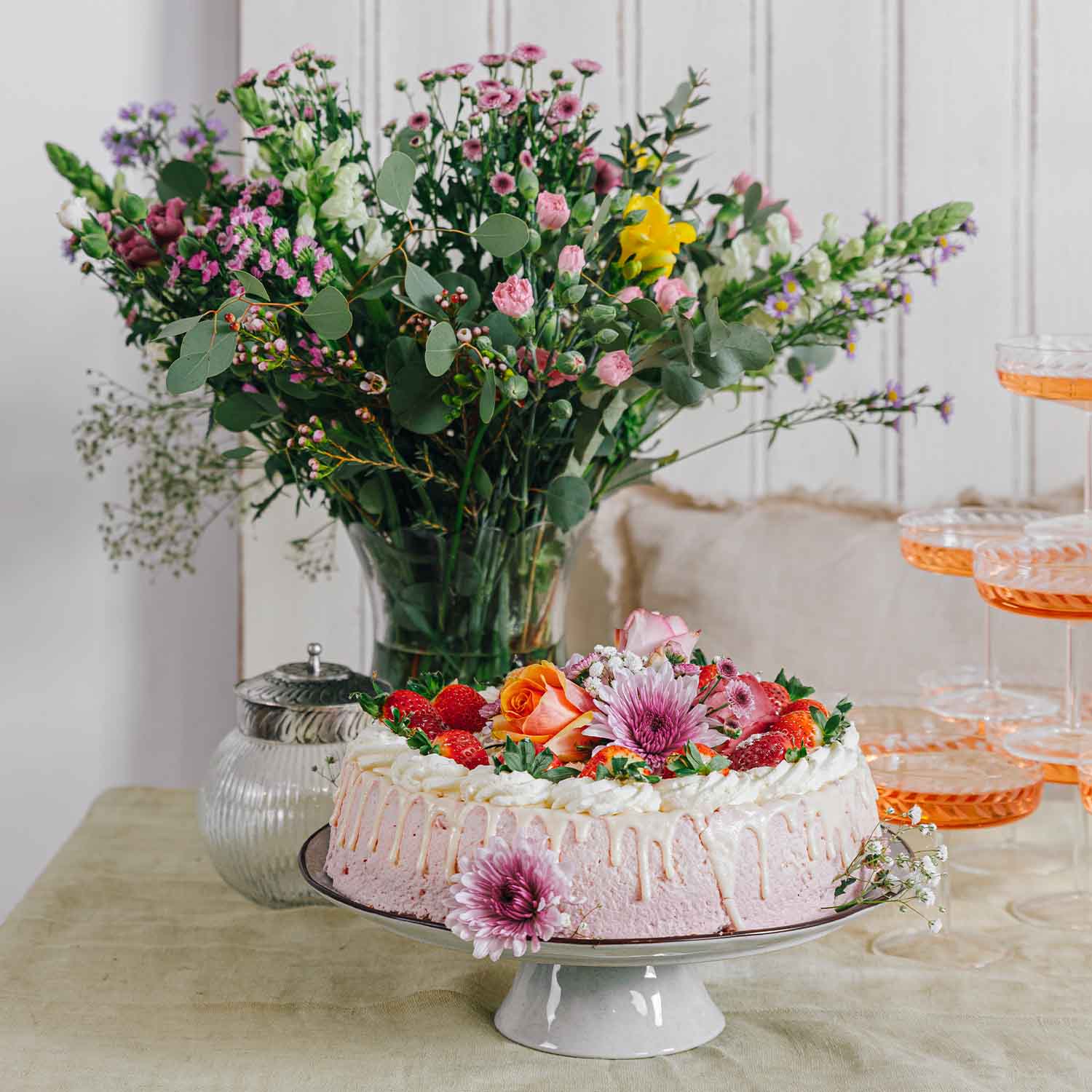 Tarta rosa con crema, fresas frescas y flores decorativas sobre soporte blanco con ramo de flores al fondo