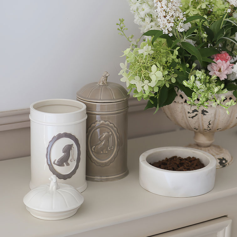 Recipientes de cerámica crema y beige para comida de perro junto a un cuenco blanco con pienso y flores