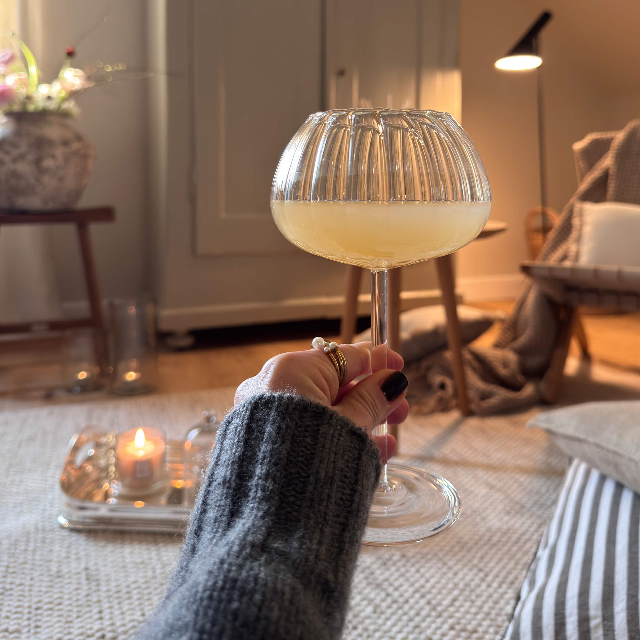 Mano sosteniendo una copa con bebida amarilla en un interior acogedor con luz suave y decoraciones