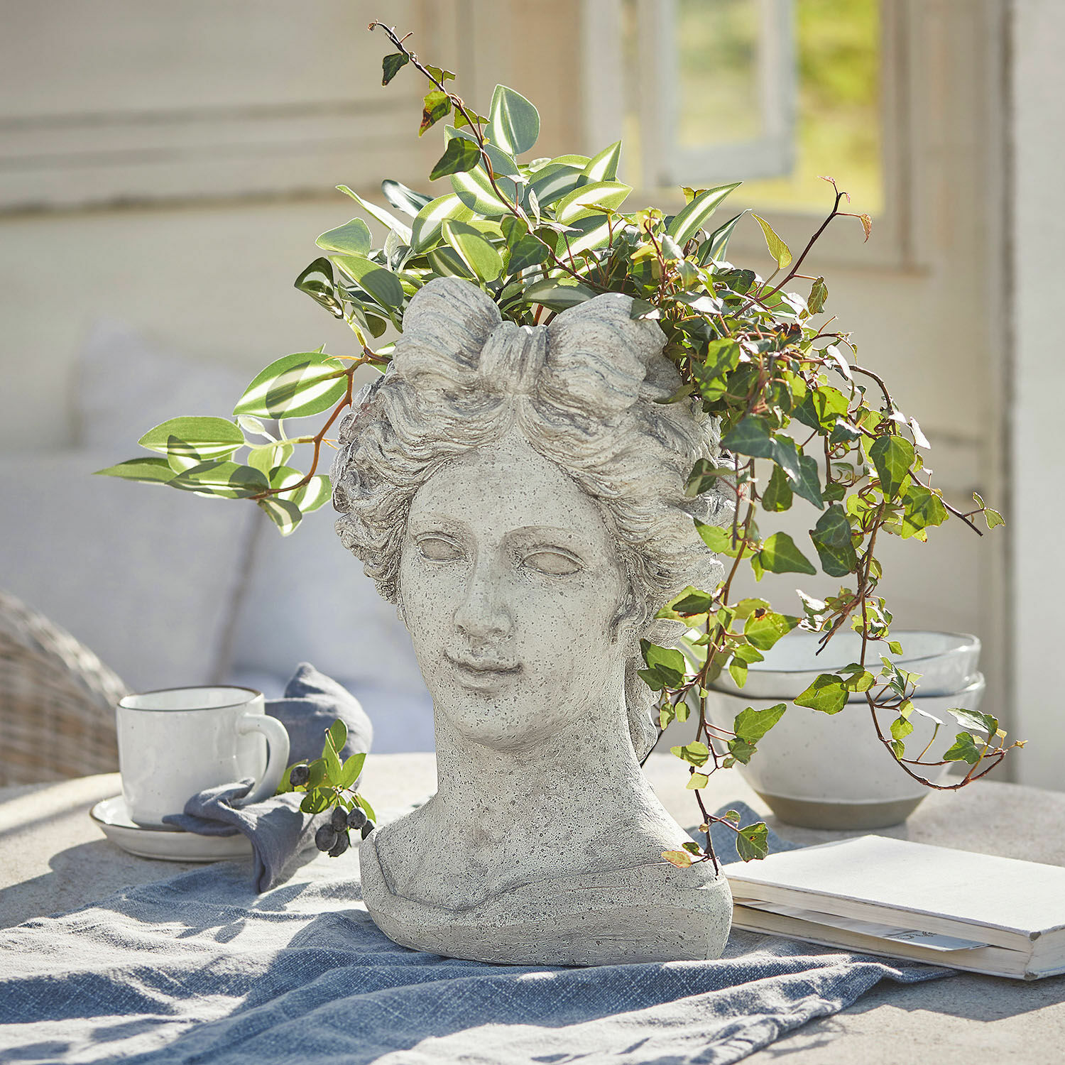 Maceta Cassiopeia Maceta en forma de cabeza de mujer de piedra gris con plantas verdes sobre mesa en interior luminoso