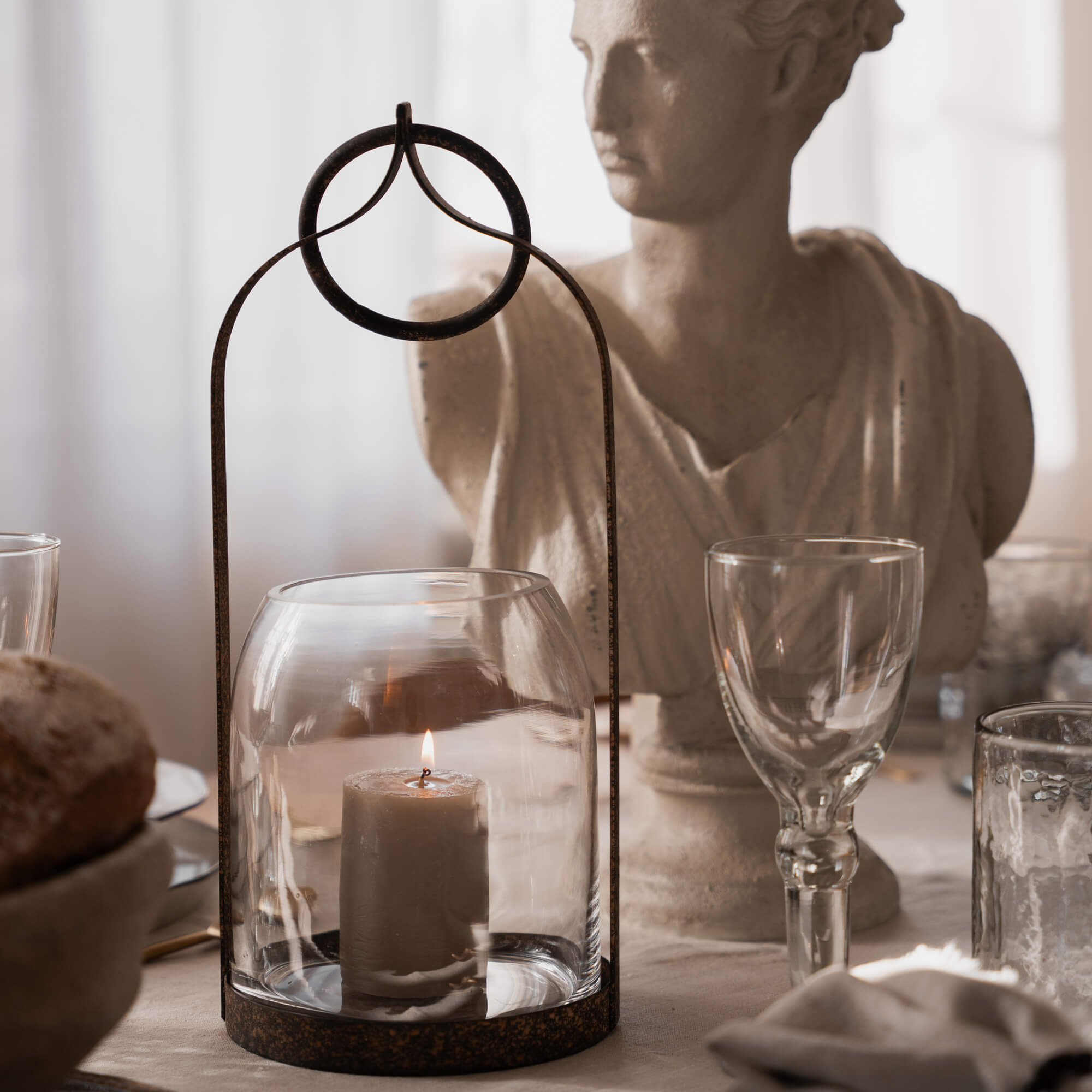 Farol de vidrio con marco metálico oxidado y vela encendida sobre mesa rústica con escultura decorativa