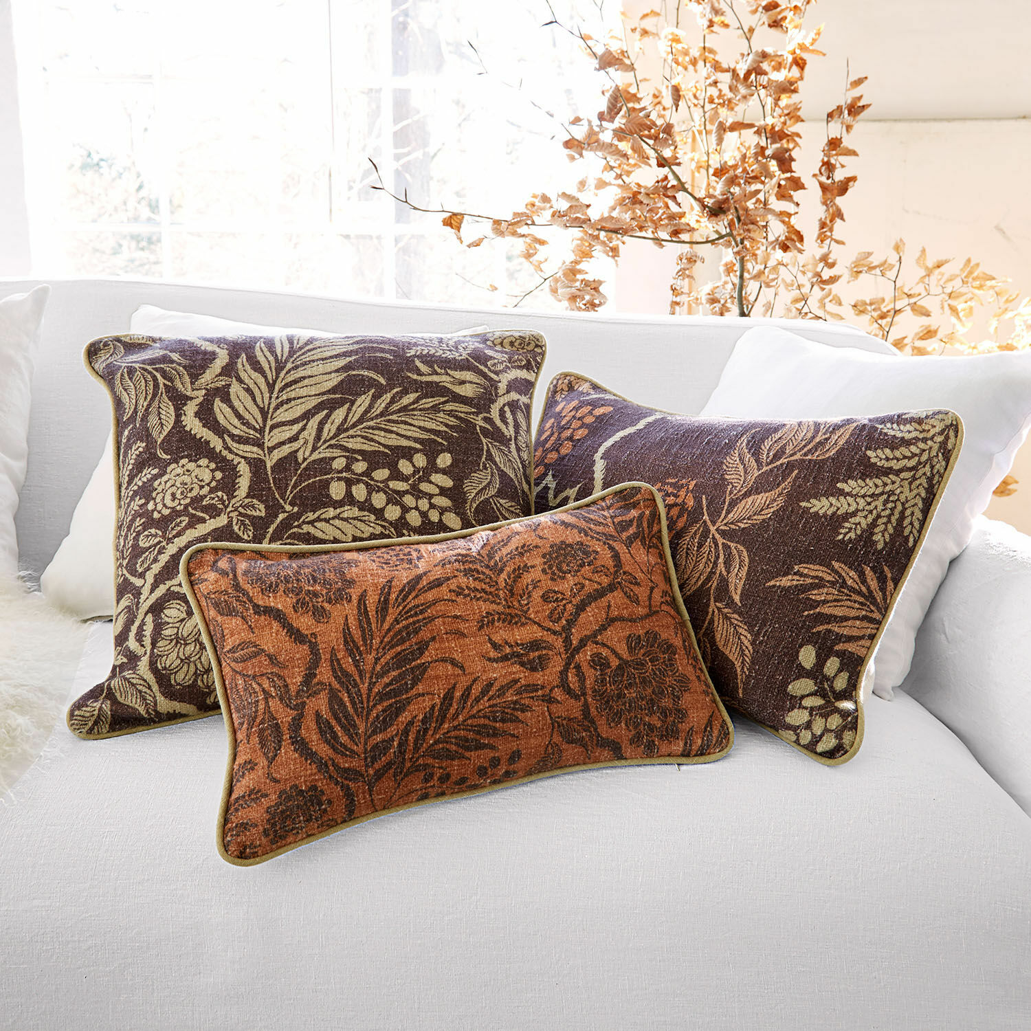 Conjunto de tres cojines decorativos con diseño de hojas en tonos marrones y naranjas sobre sofá blanco