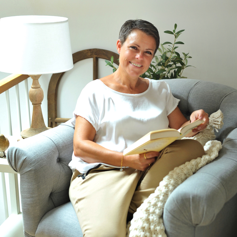 Mujer con camiseta blanca y pantalones beige leyendo un libro en un sofá gris cómodo en una habitación luminosa
