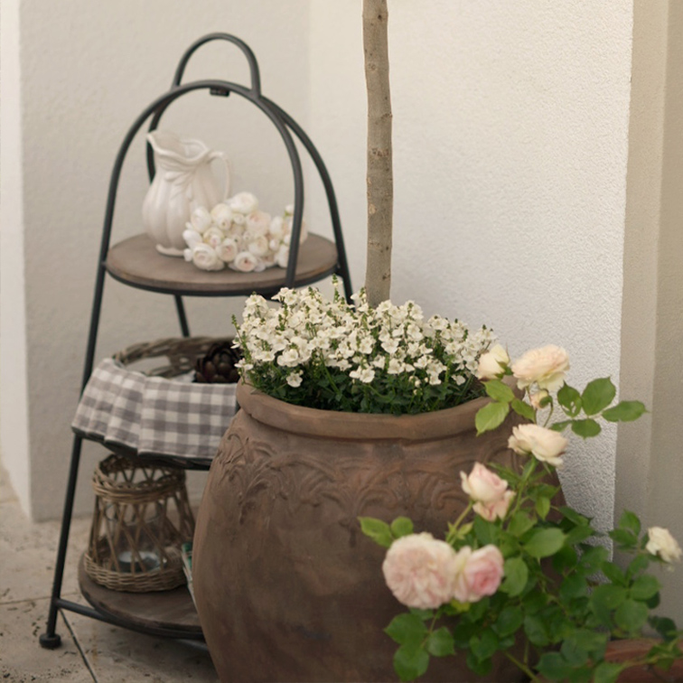 Maceta grande de cerámica con flores blancas y rosas junto a estantería metálica con jarra y farol