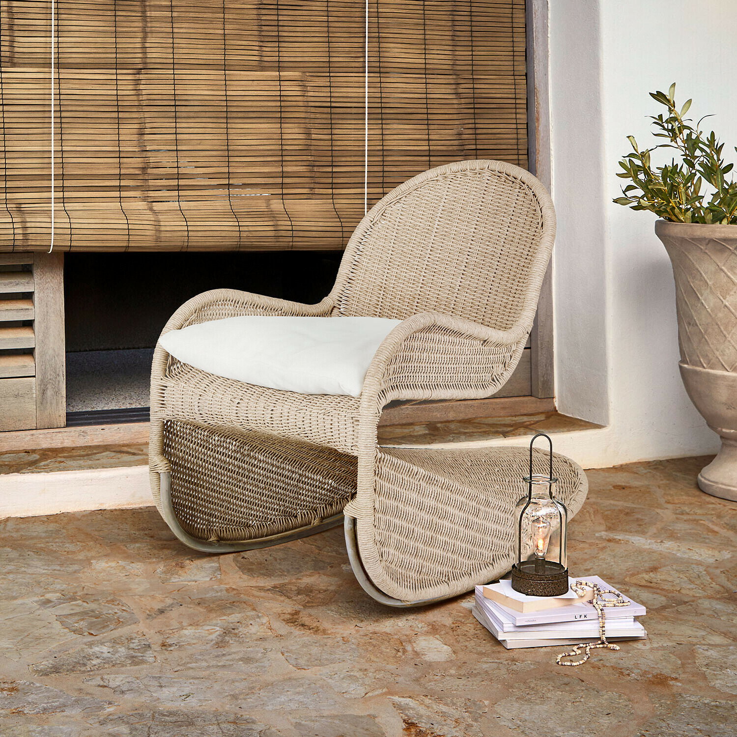 Mecedora Fernleigha Sillón de ratán beige con cojín blanco sobre suelo de piedra junto a persiana de bambú
