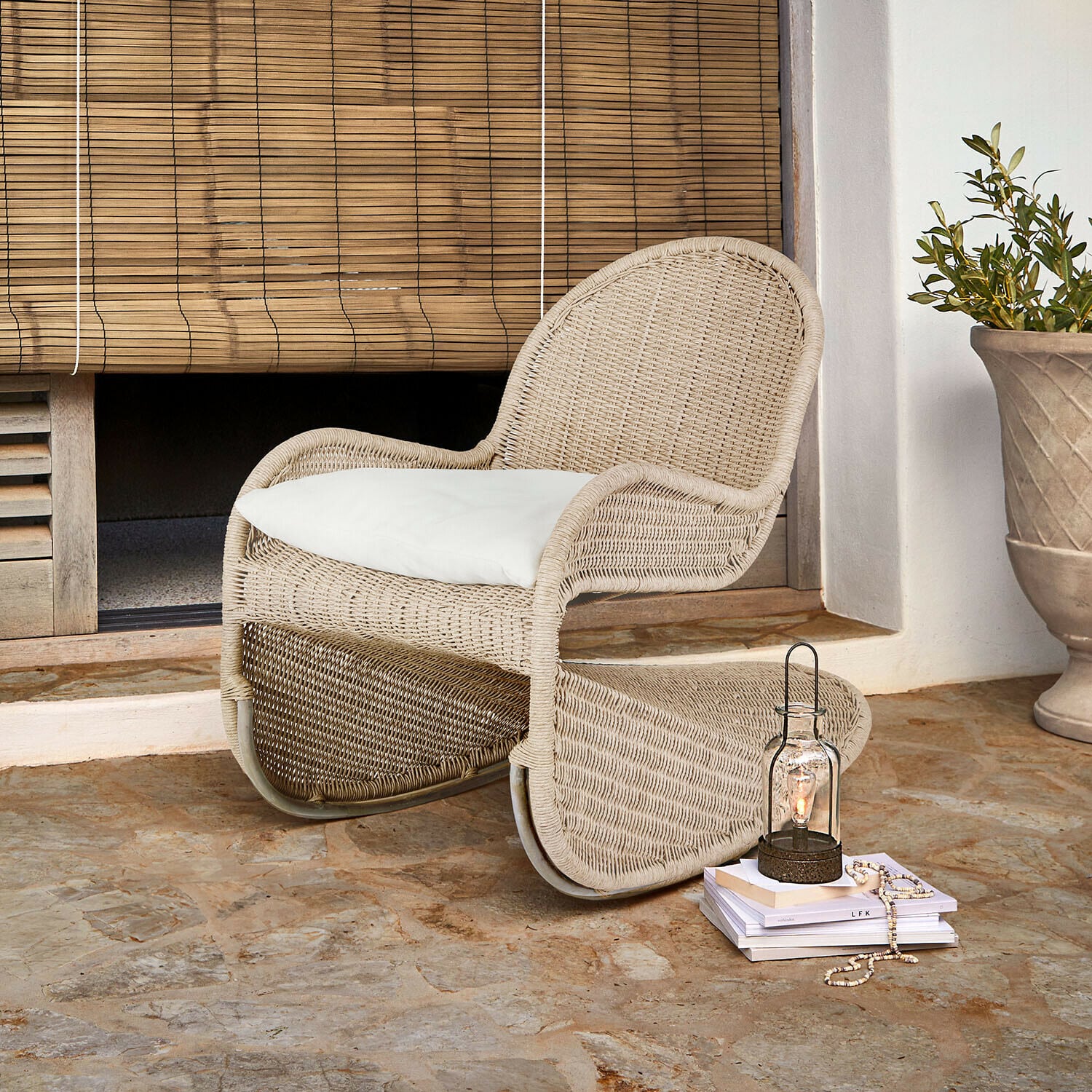 Mecedora Fernleigha Sillón de ratán beige con cojín blanco sobre suelo de piedra junto a persiana de bambú