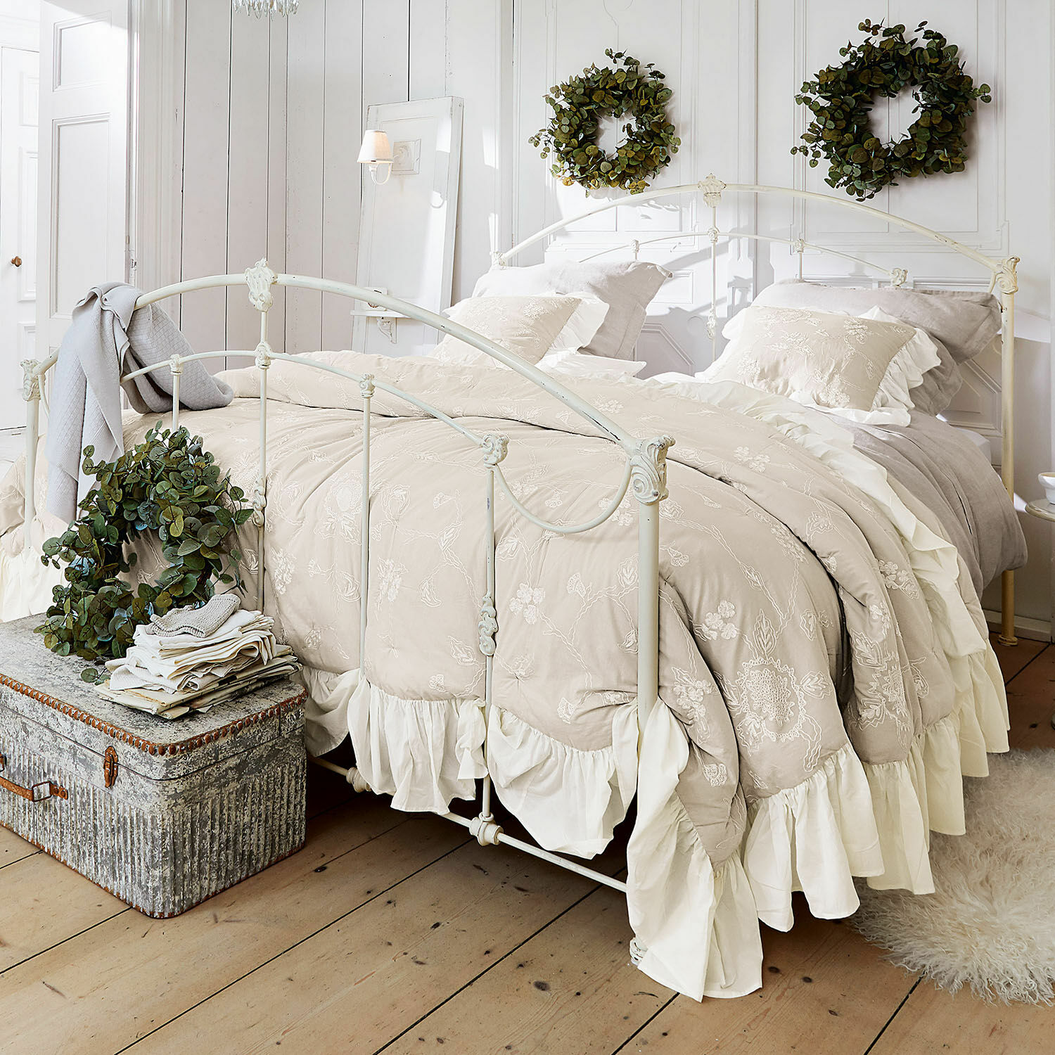 Colcha Clarissa Ropa de cama beige bordada con volantes en cama metálica blanca en dormitorio luminoso con coronas verdes