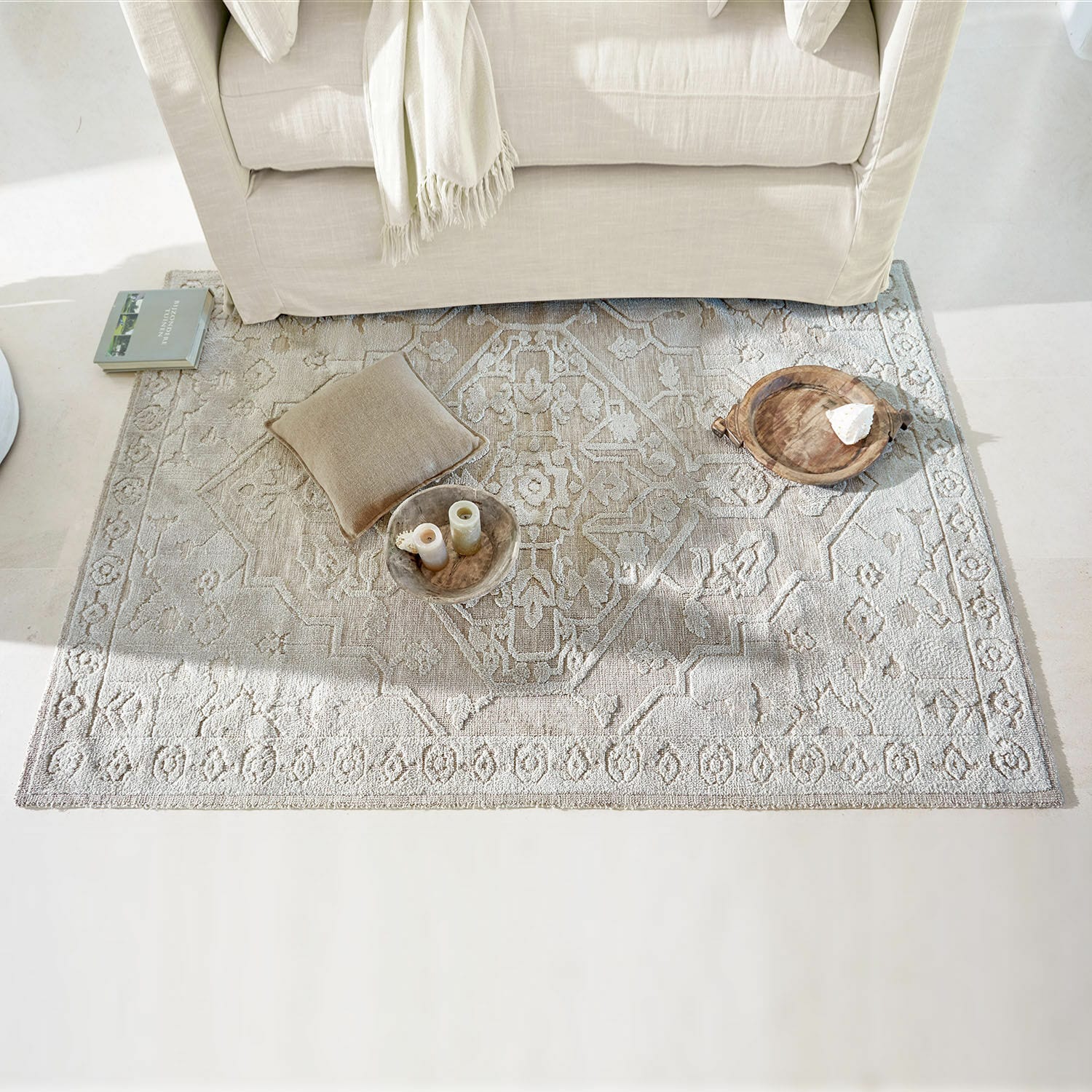 Alfombra Armidale Alfombra beige con diseño bajo un sofá claro con cojín y decoraciones de madera en sala luminosa