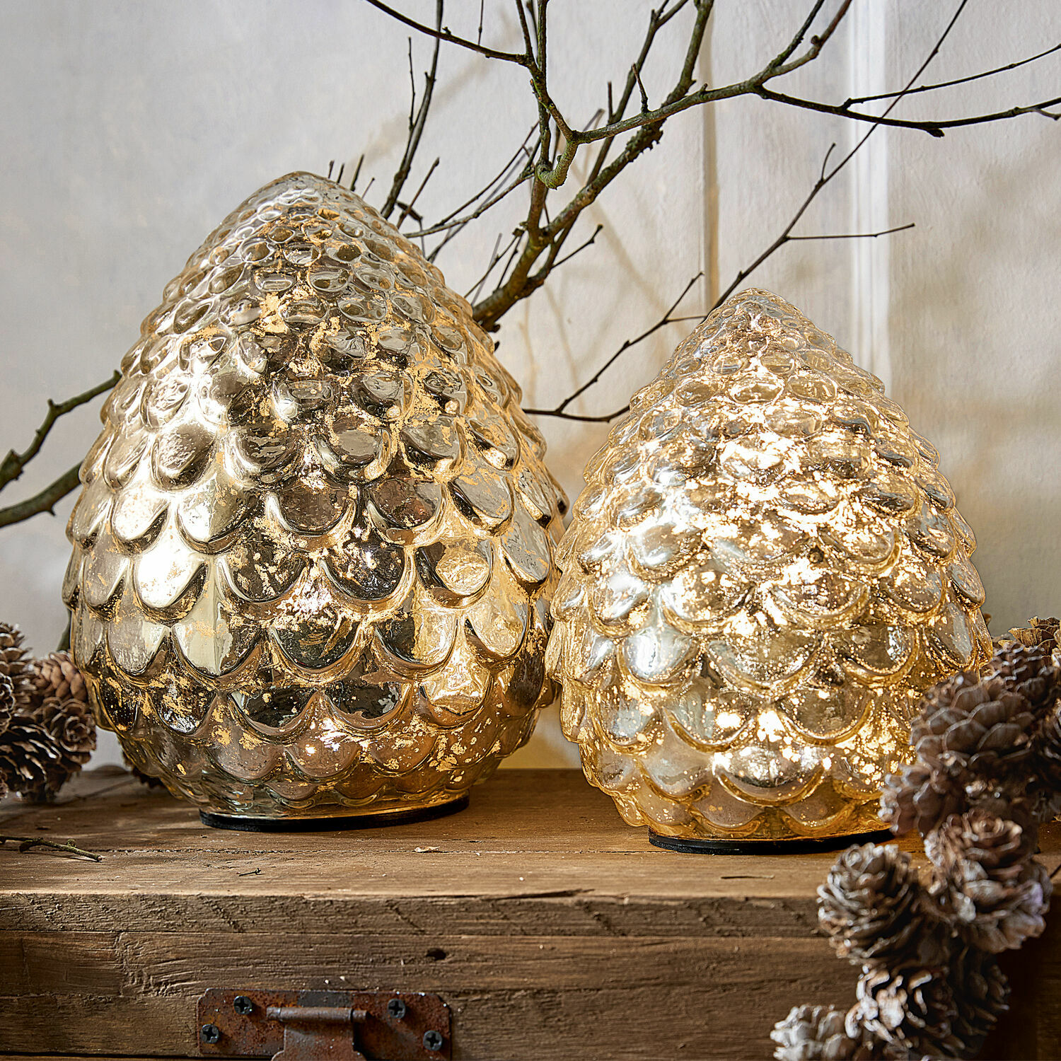 Set de 2 conos LED Ibaque Dos conos decorativos de vidrio dorado con textura de escamas sobre superficie de madera con piñas