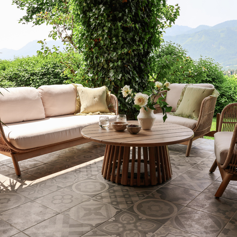 Muebles de jardín de ratán con cojines beige y mesa redonda de madera en terraza con baldosas decorativas