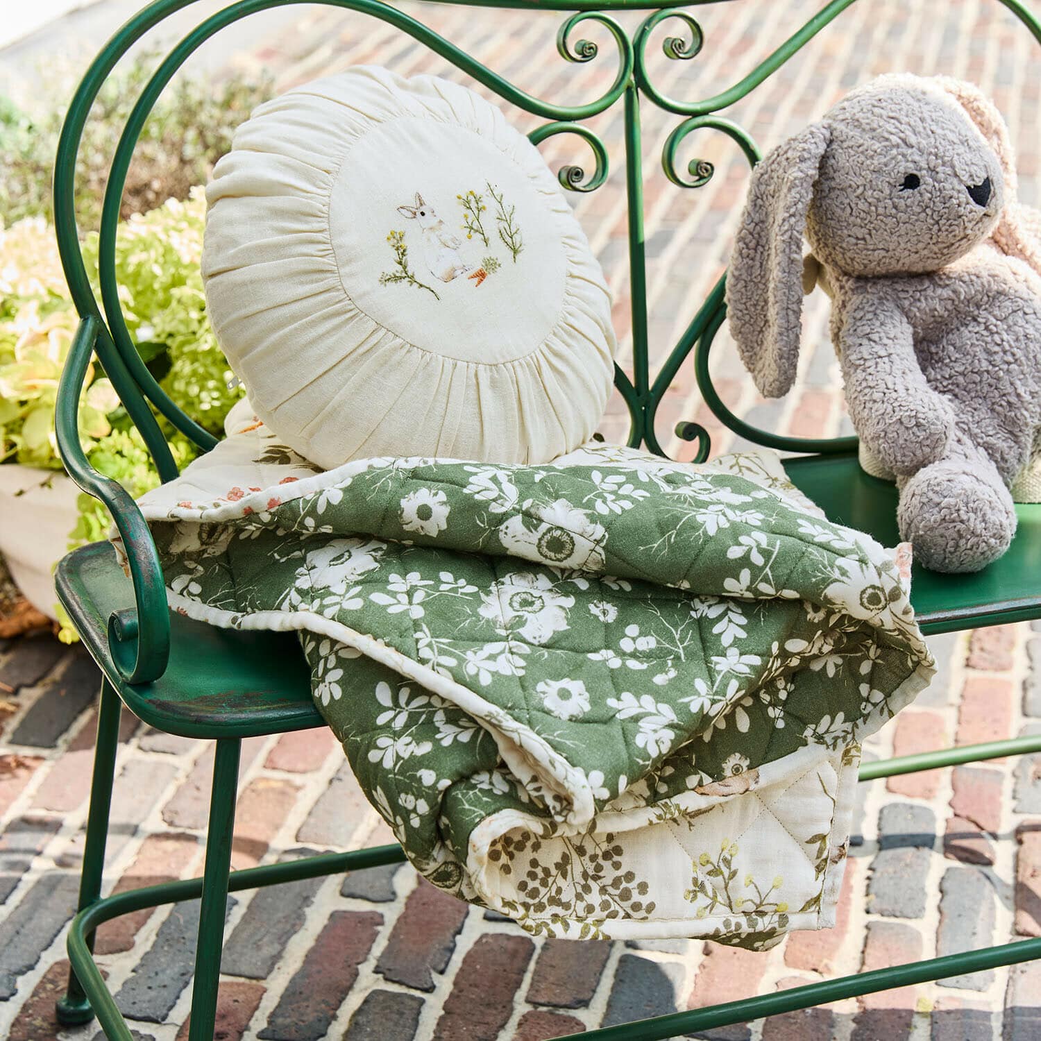 Plaid y cojín Puddlebrook Manta acolchada verde con estampado floral blanco sobre banco metálico verde con cojín redondo y peluche de conejo