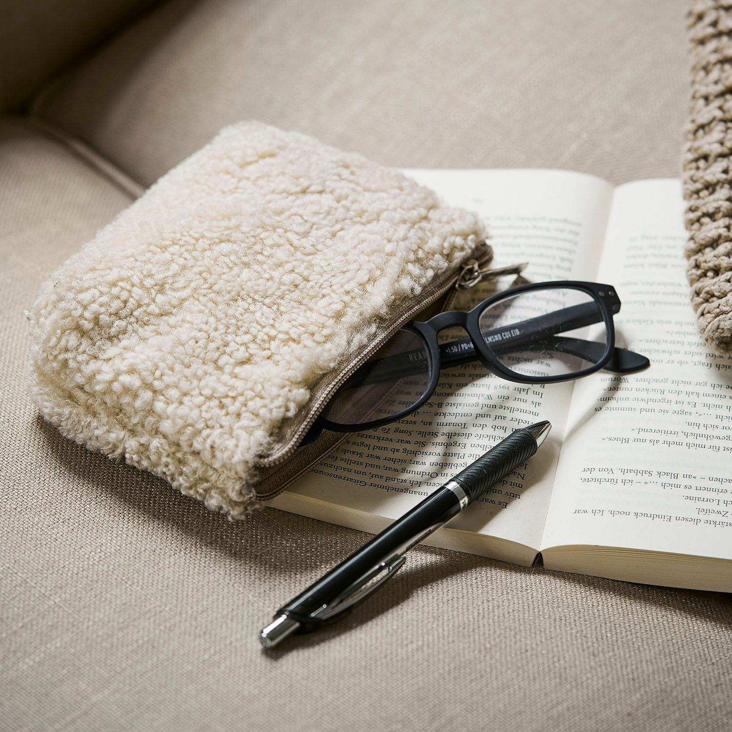 Bolsita Bathilde Neceser beige de material tipo borrego sobre sofá con gafas y bolígrafo negro