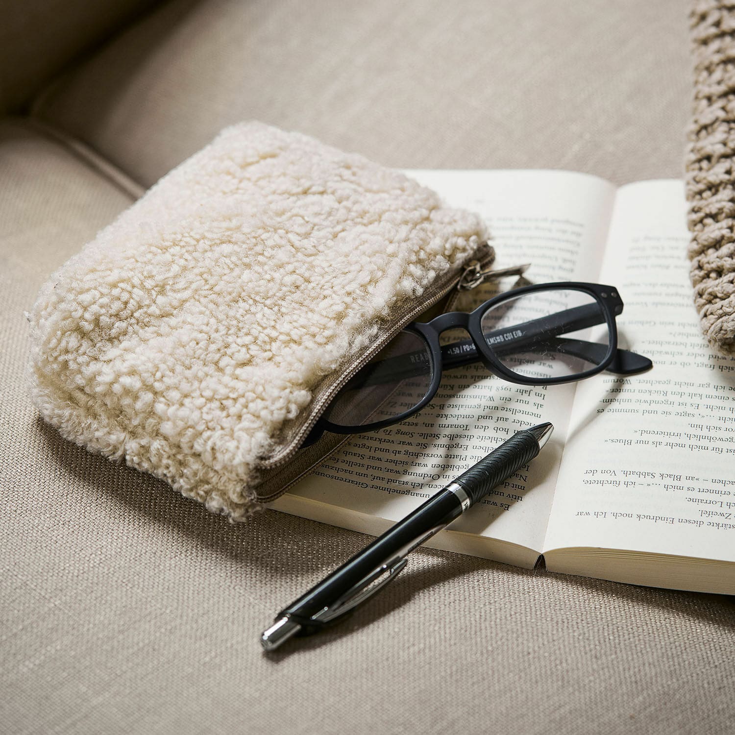 Bolsita Bathilde Neceser beige de material tipo borrego sobre sofá con gafas y bolígrafo negro