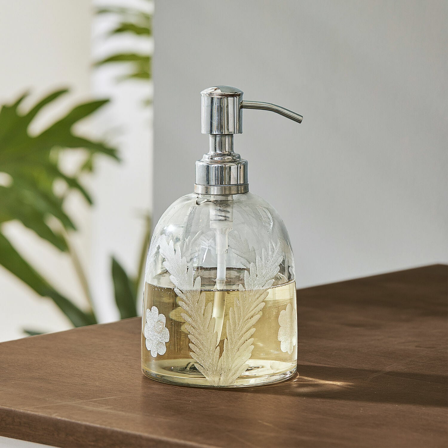 Dispensador de jabón Crystal Dispensador de jabón de vidrio con bomba metálica y diseño de hojas blancas sobre mesa marrón