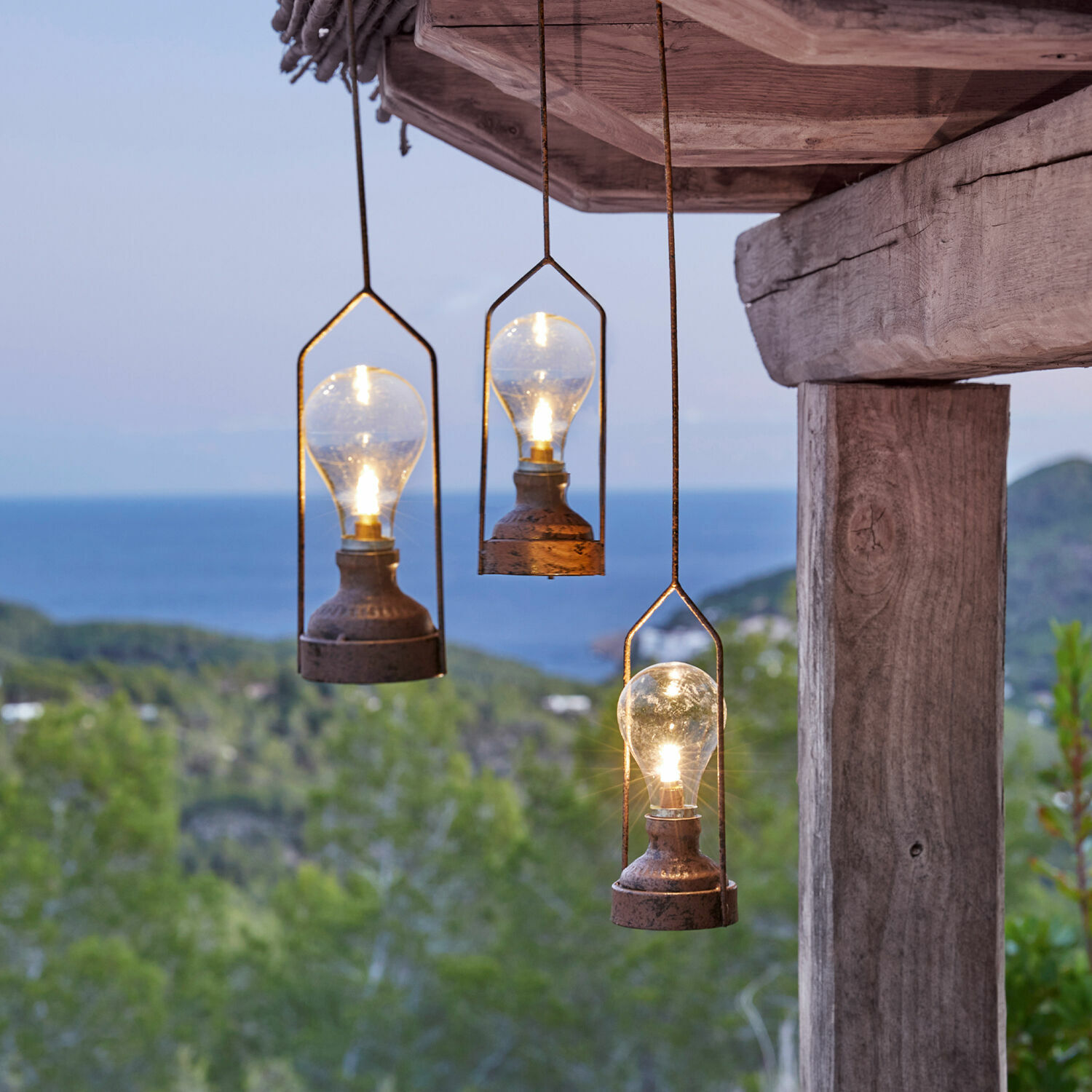 Set de 3 farolillos colgantes LED Chambéry Tres lámparas colgantes de metal con bombillas frente a bosque verde y mar, iluminación exterior