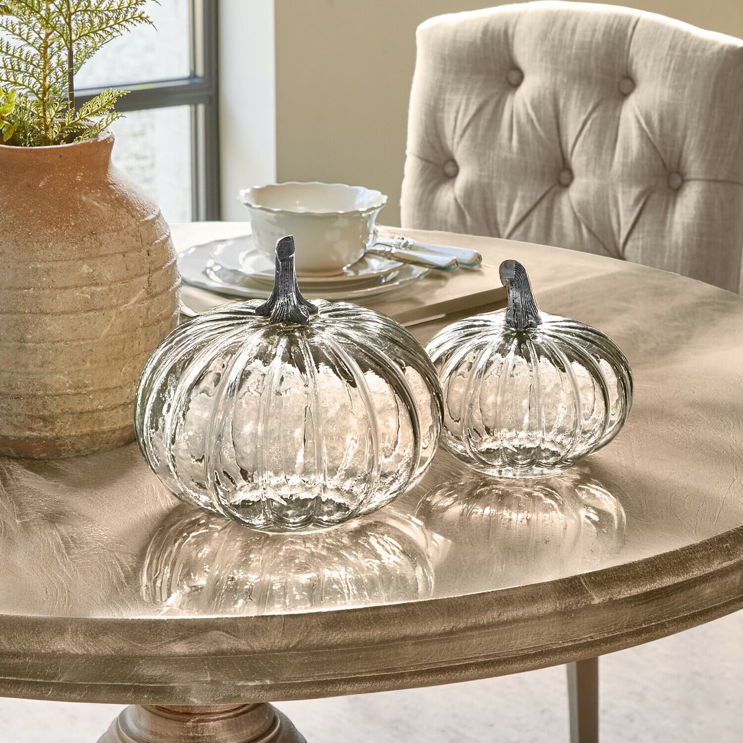 Set de 2 calabazas Perlouin Dos calabazas de cristal transparente con tallos metálicos sobre una mesa decorativa beige