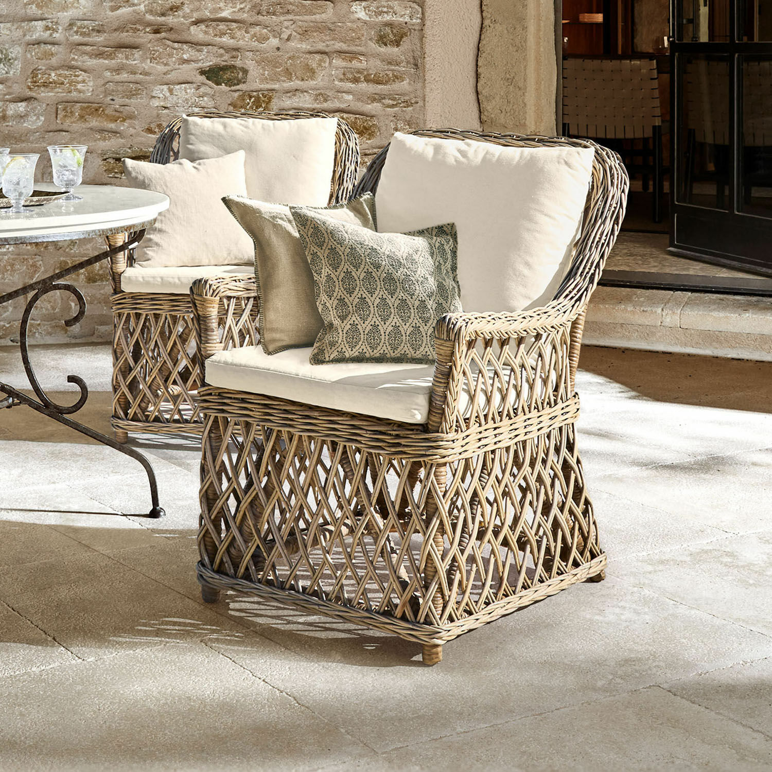 Sillón Nesvryn Sillón cómodo de ratán natural con cojines crema y cojines decorativos para terraza elegante.